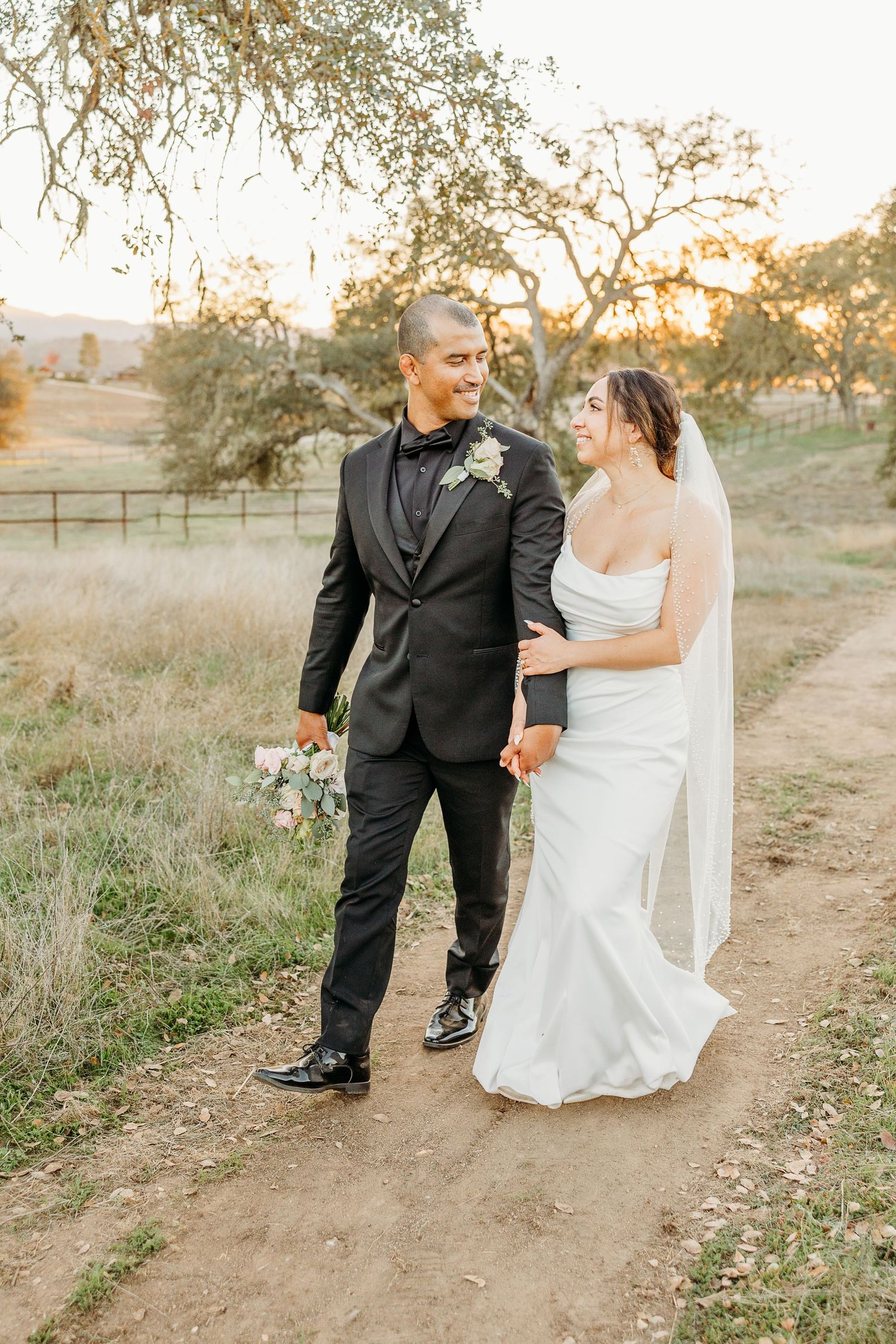 A couple in wedding attire strolls along a rustic dirt path at sunset.