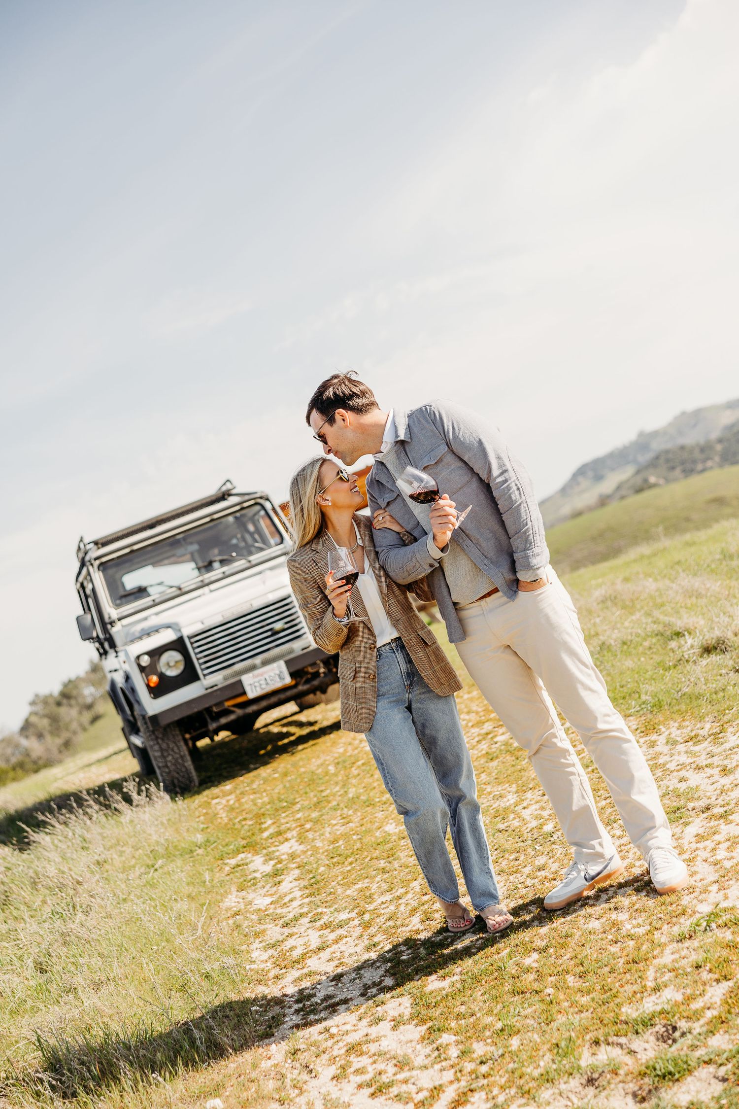A couple stands in a sunny field with a vintage truck in the background during a romantic countryside photoshoot.