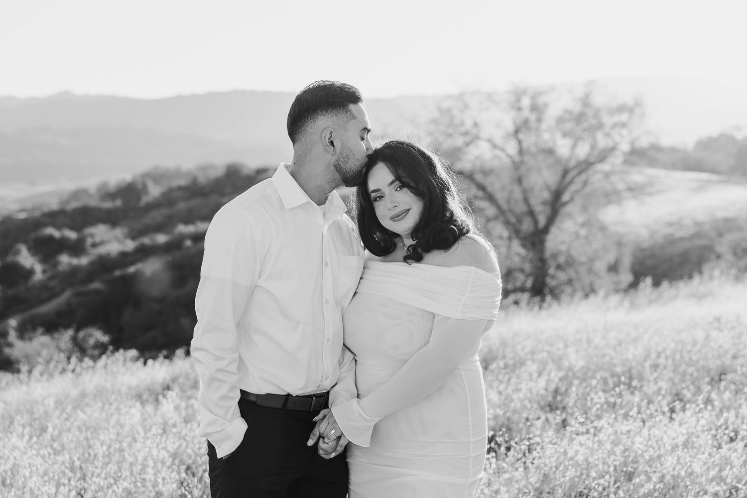 A couple shares an intimate moment in a scenic field of white wildflowers with rolling hills in the background.