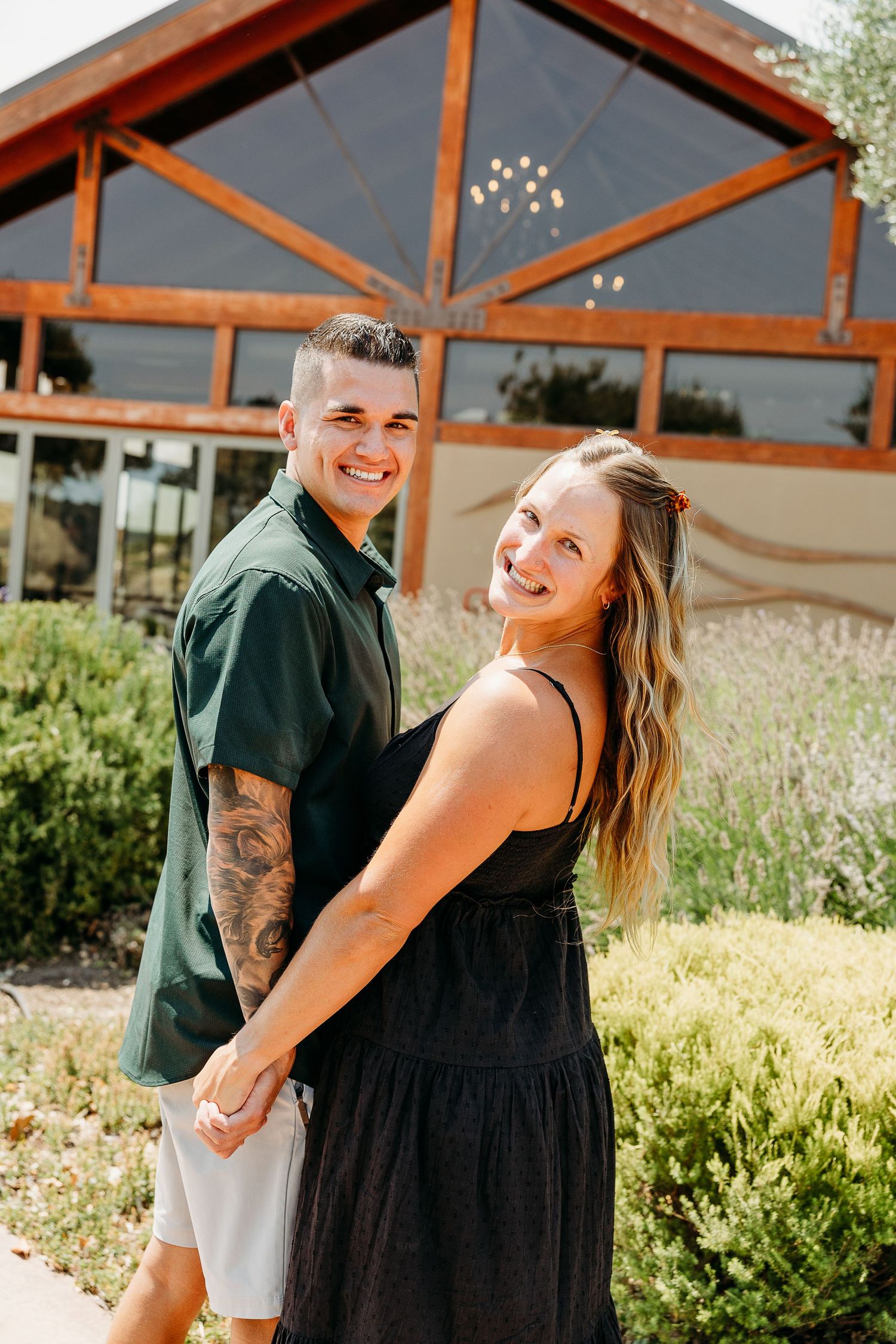A couple shares a romantic moment in front of rustic wooden beams and greenery during an outdoor photoshoot.