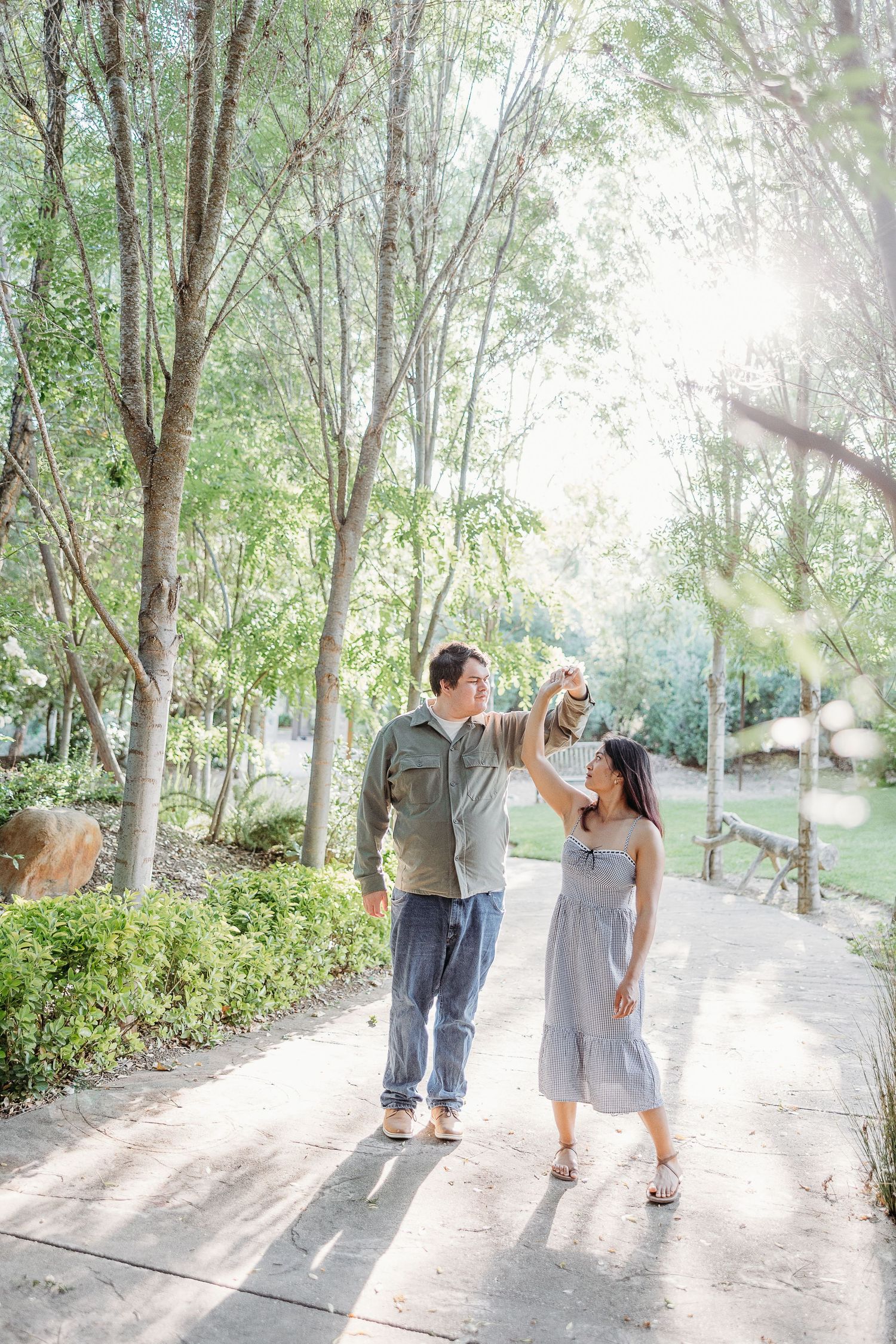 A couple walking together along a sunlit tree-lined pathway in a garden setting on a bright sunny day.