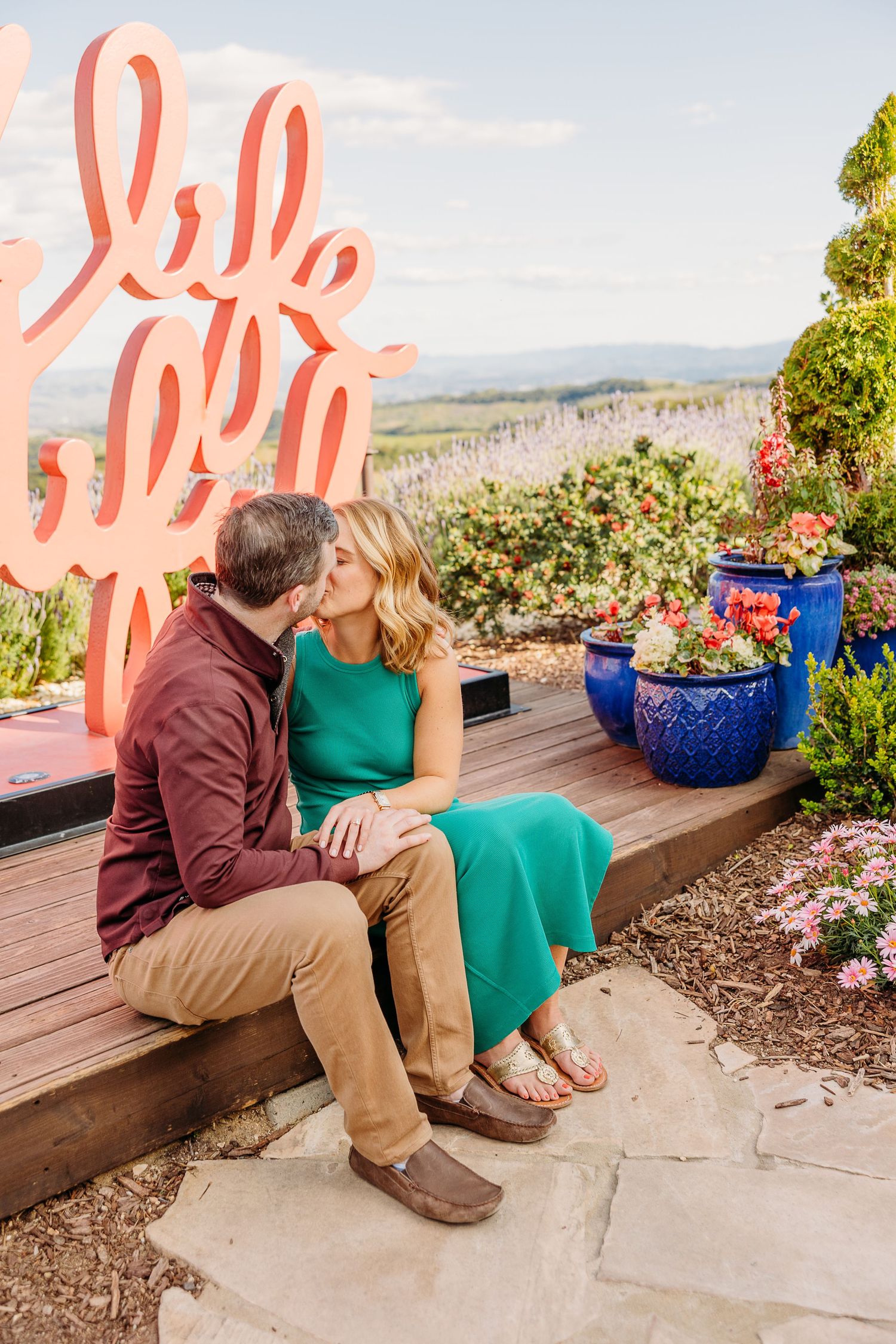 Engaged couple sits on outdoor deck with mountain views and vibrant flower pots in the scenic background.