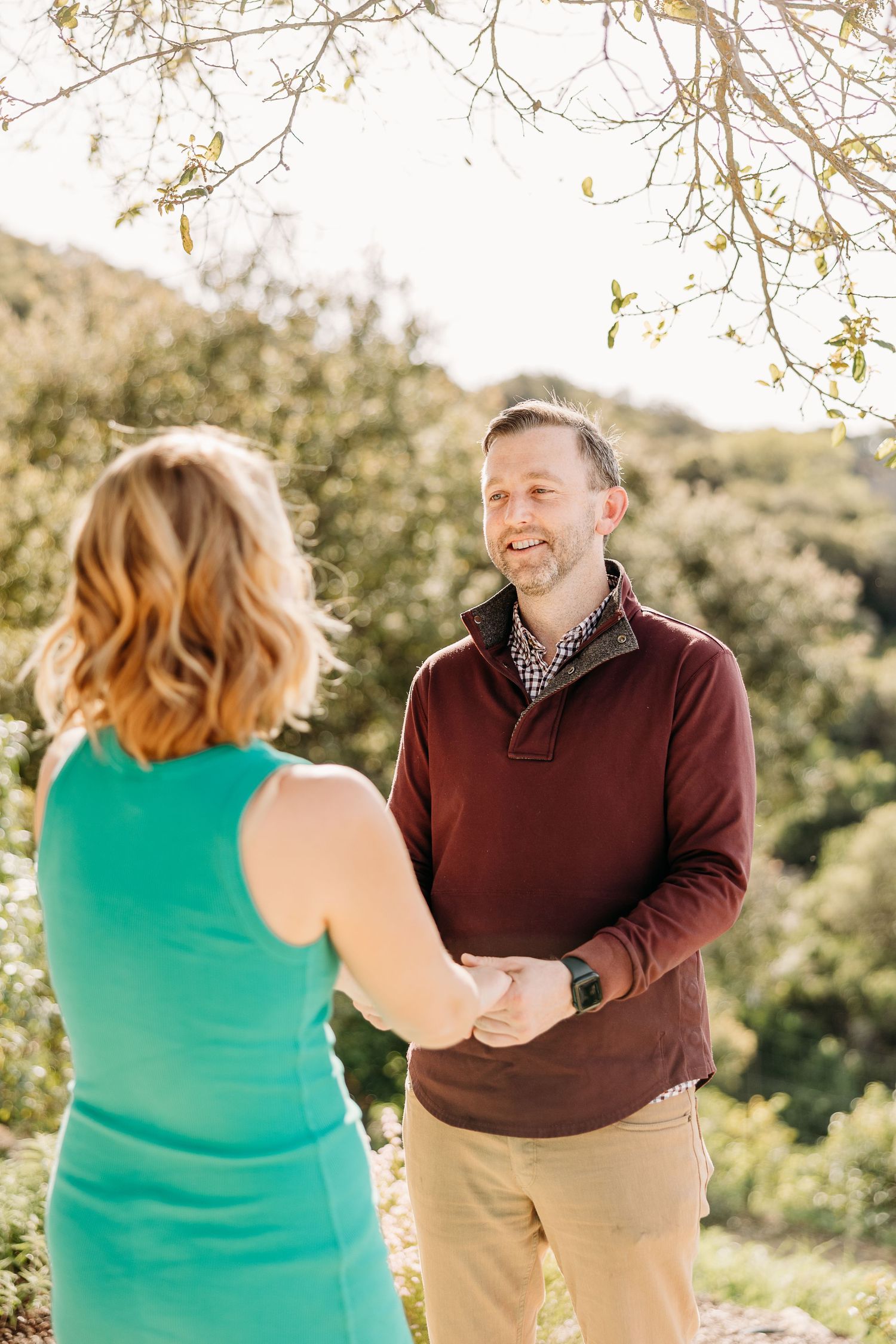 A couple shares joyful laughter together in a sunlit outdoor setting with natural greenery in the background.