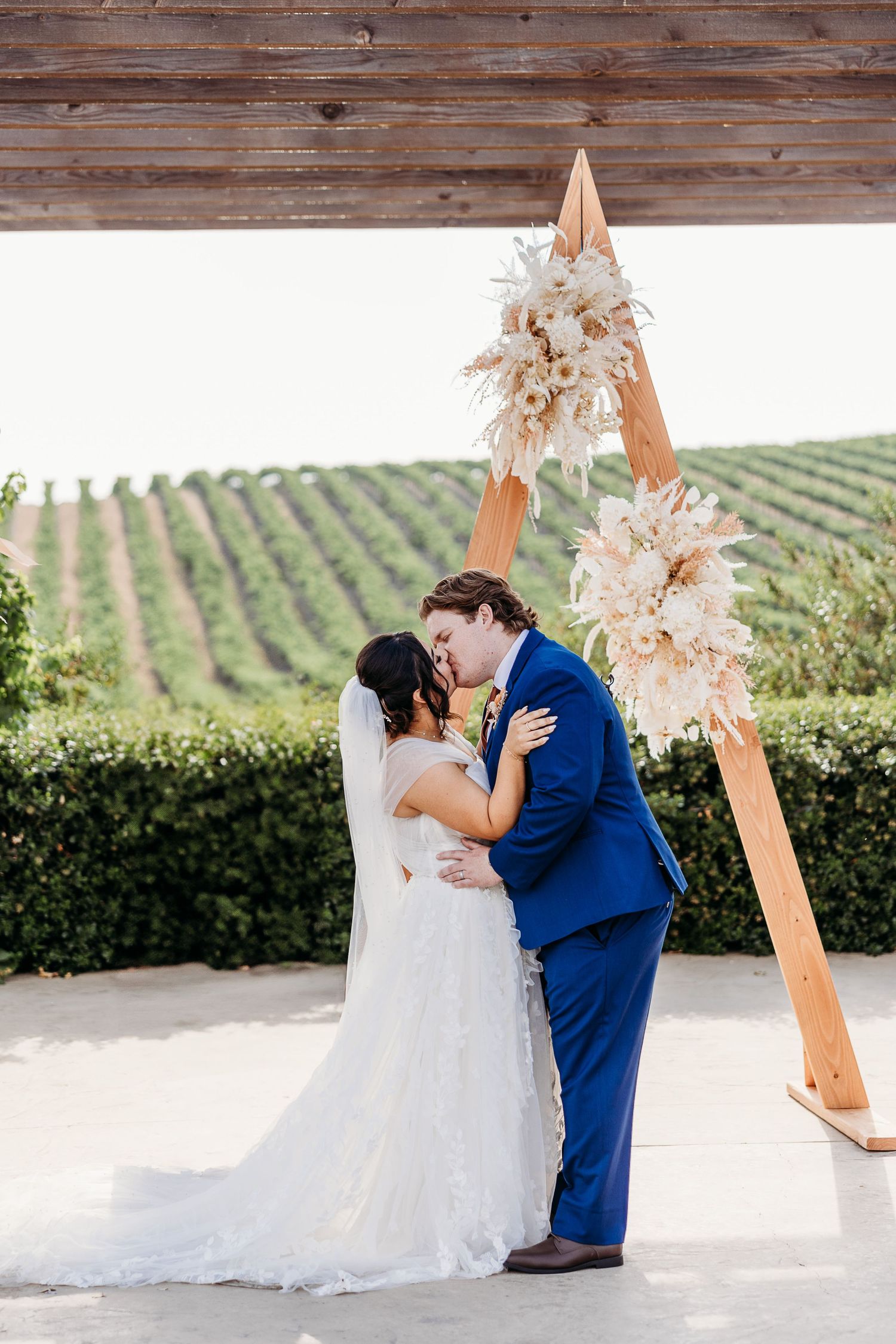 A couple shares their first kiss under a wooden arch decorated with dried flowers at a vineyard wedding venue.