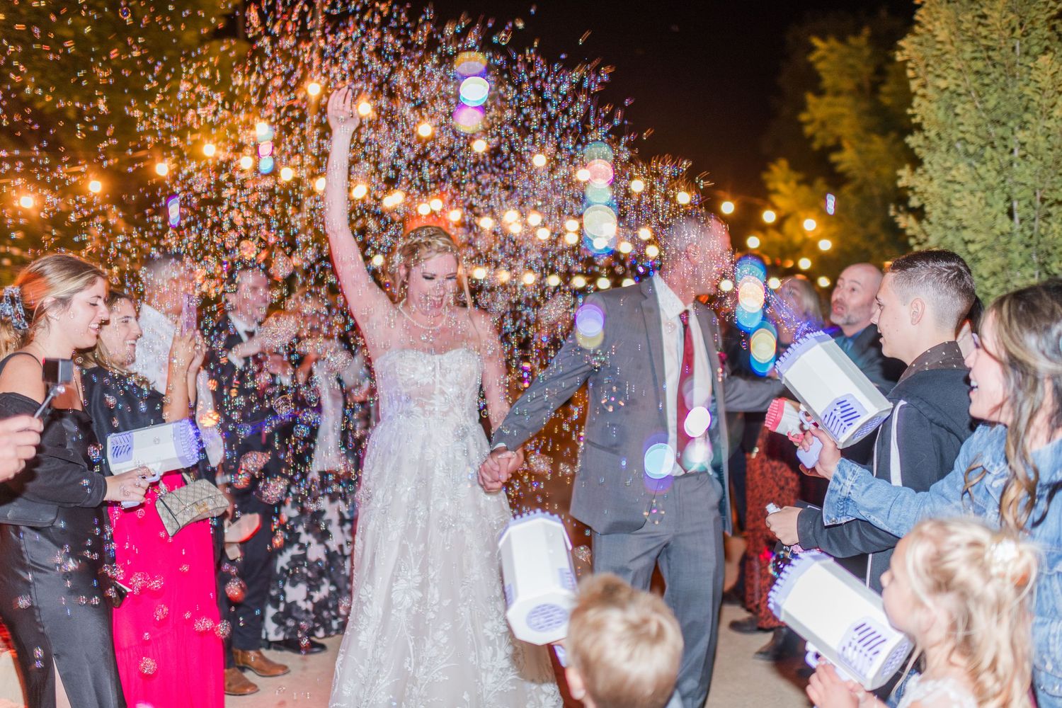 Newlyweds celebrate under falling confetti during nighttime reception with string lights.