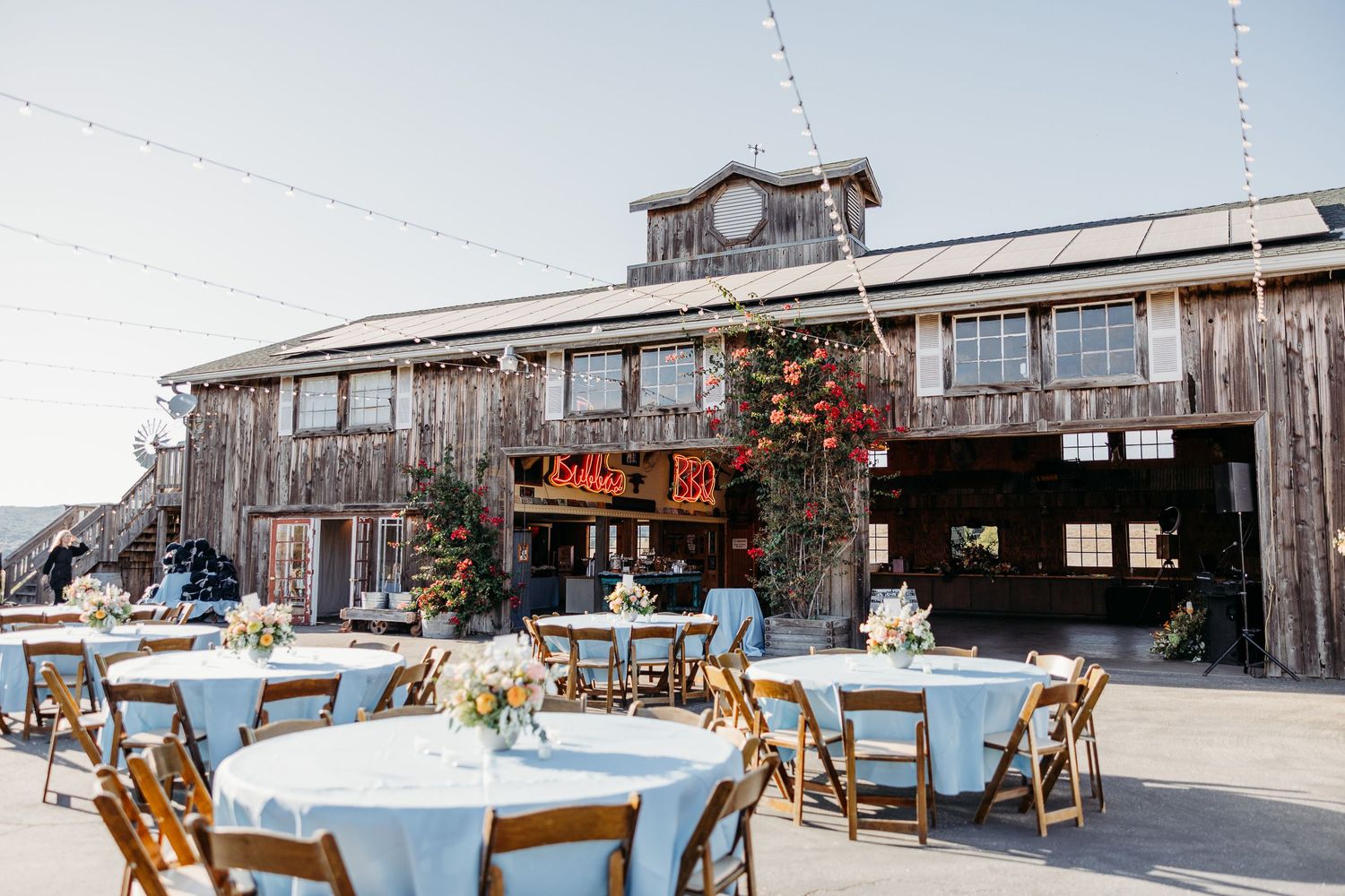 A rustic wooden barn venue with outdoor reception tables decorated with light blue linens and wooden folding chairs.