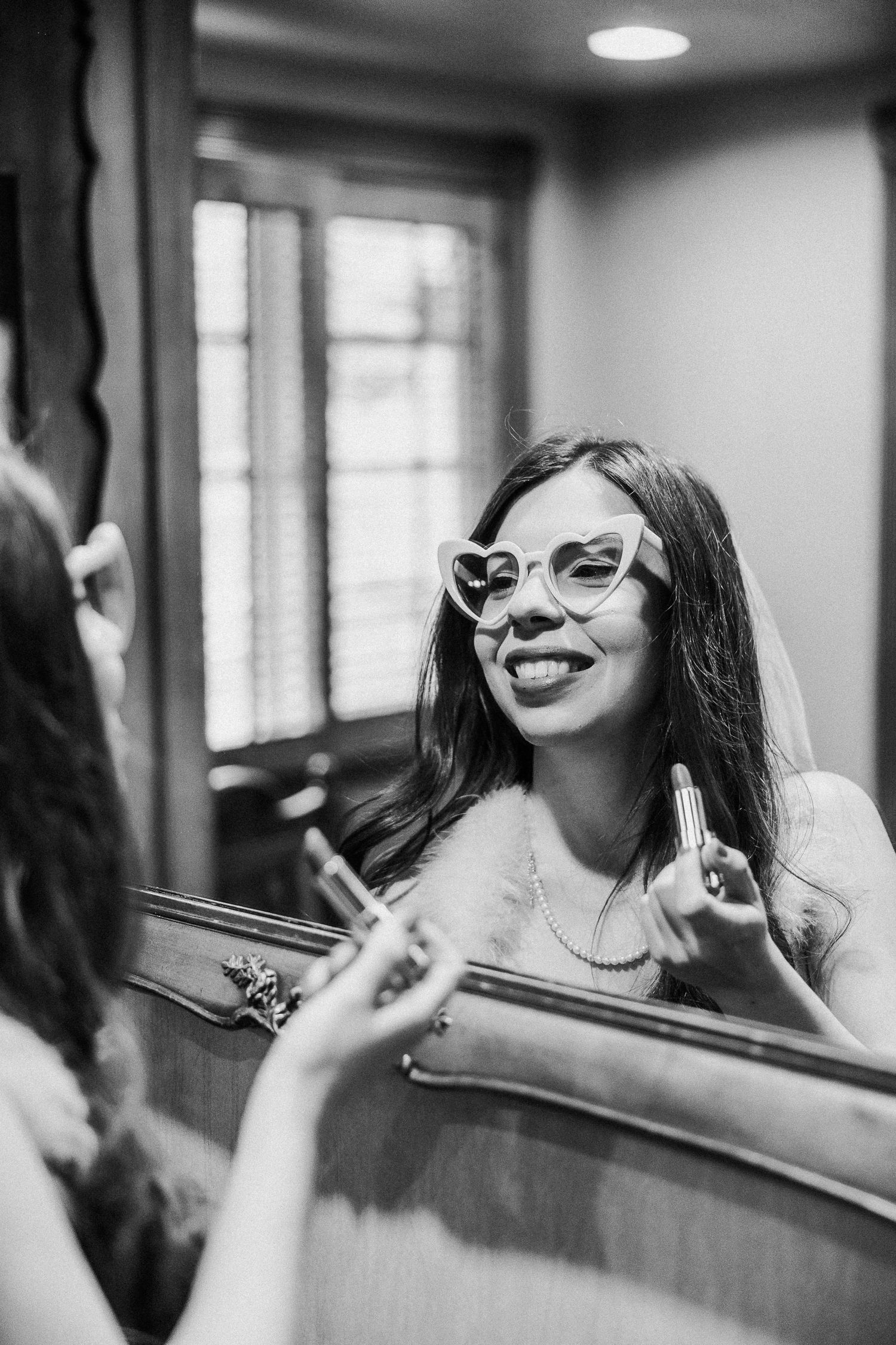 Black and white photo showing someone with long dark hair and cat-eye glasses smiling while looking in an ornate mirror.