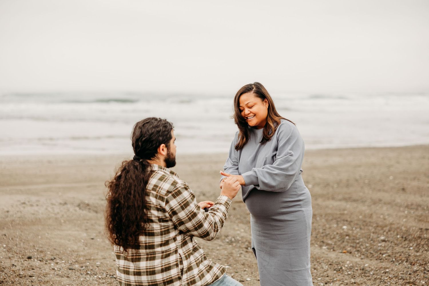 A proposal scene on a misty beach with a person in plaid kneeling before someone in a gray dress.