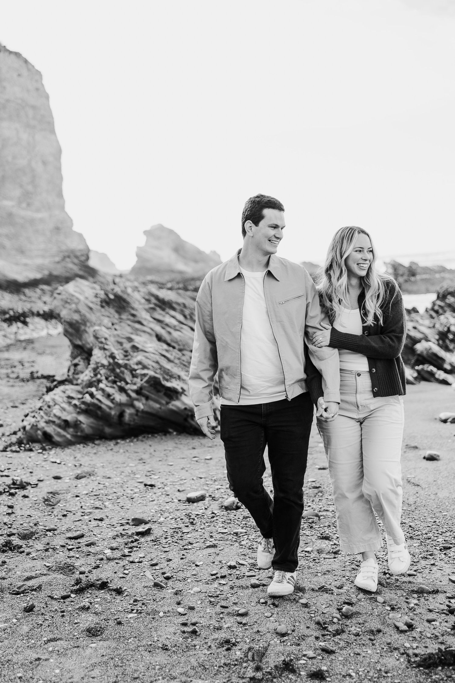 A couple walks together along a rocky beach with dramatic coastal cliffs in the background in black and white.