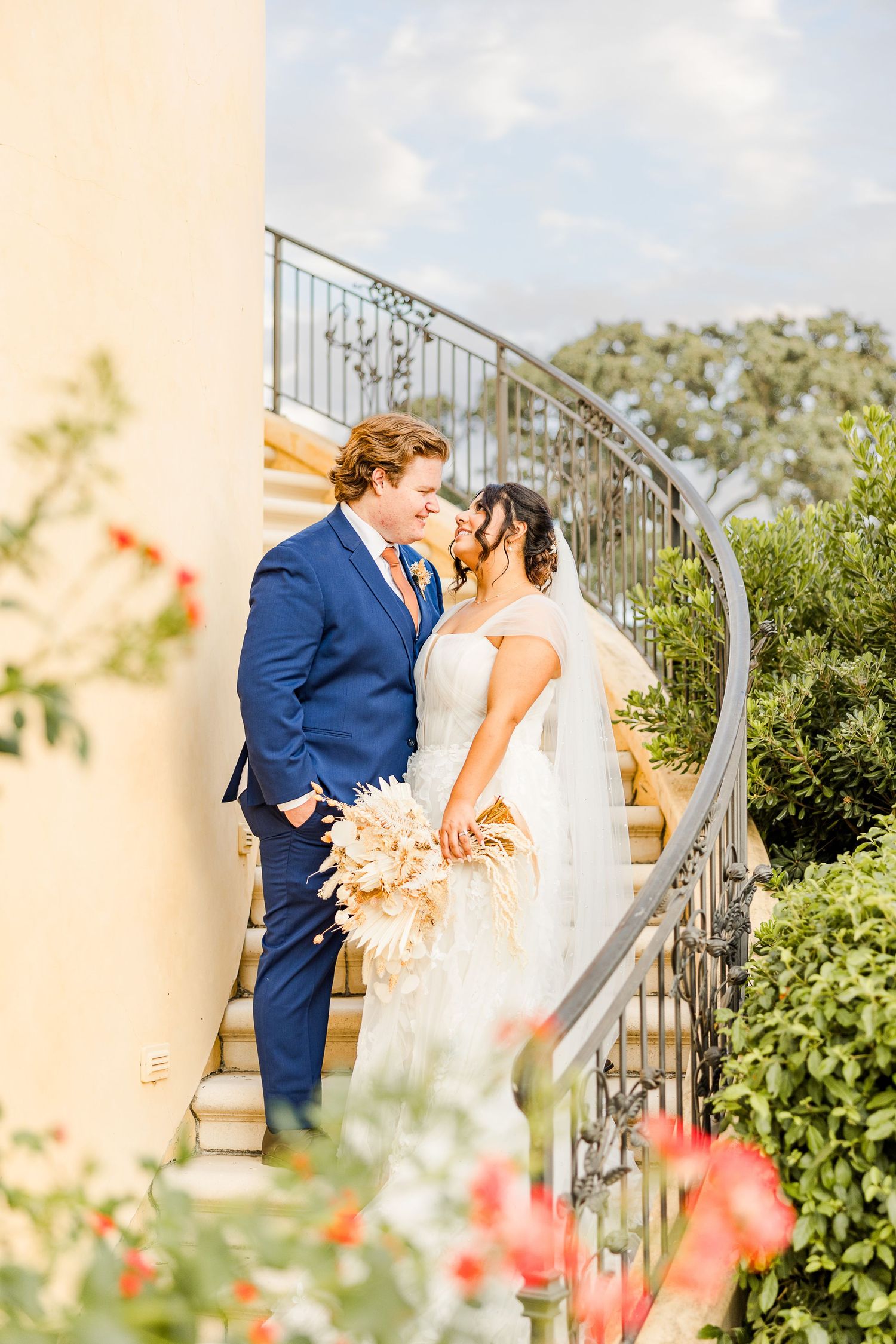 Newlyweds share a romantic moment on elegant curved stairs surrounded by blooming flowers.