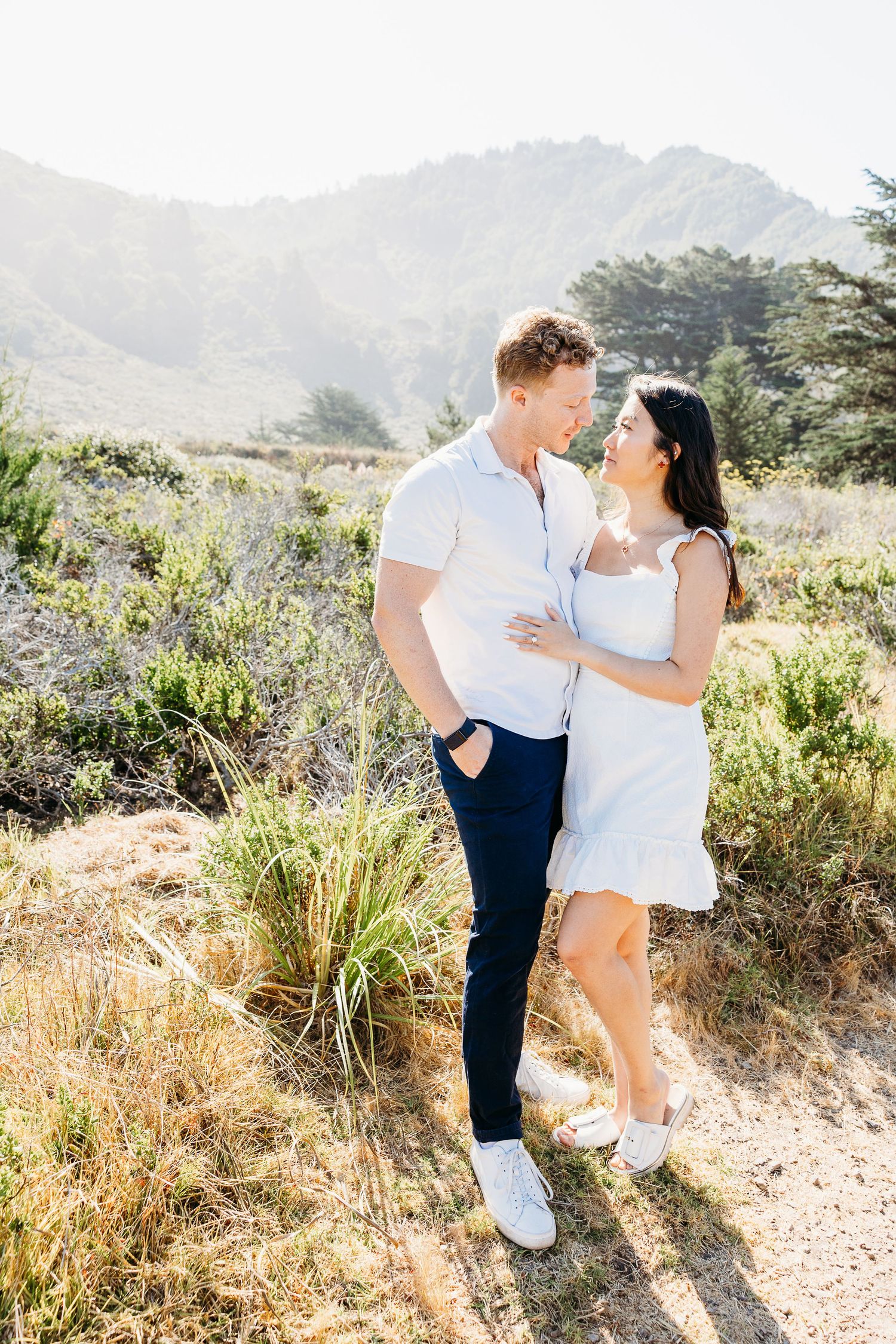 Stylish couple in white and navy outfits pose on scenic mountain trail.