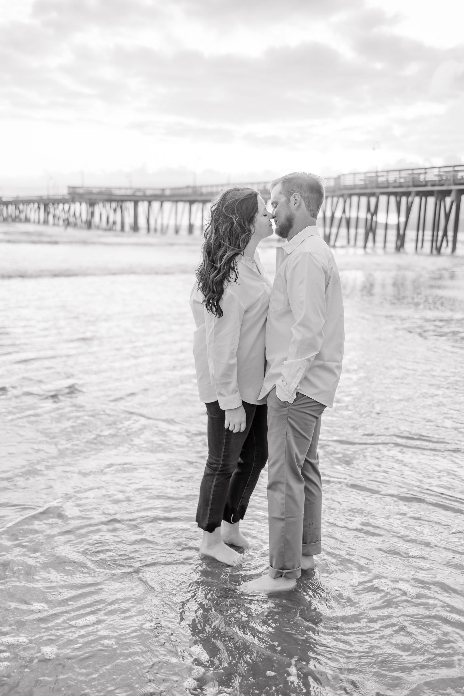 Black and white photo of a couple standing in shallow ocean water near a long wooden pier.