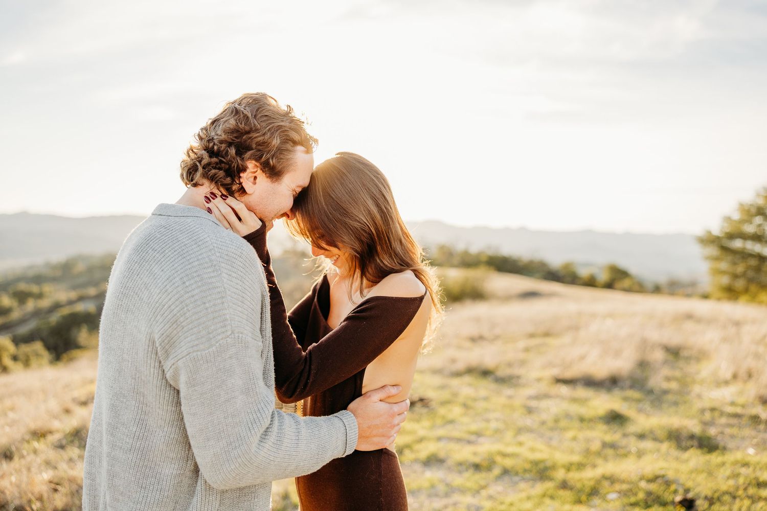 Couple embracing tenderly at golden hour in a scenic field with rolling hills in the background.