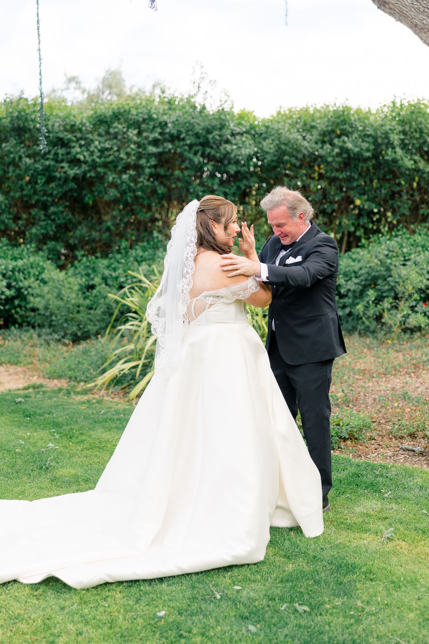 Bride in white gown and veil shares emotional first look moment with older man in black suit on green lawn.