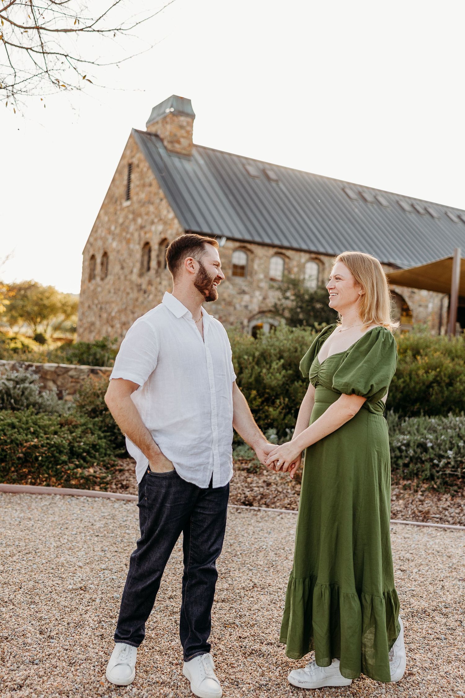 Couple in white shirt and green dress posing playfully outside stone building at golden hour.