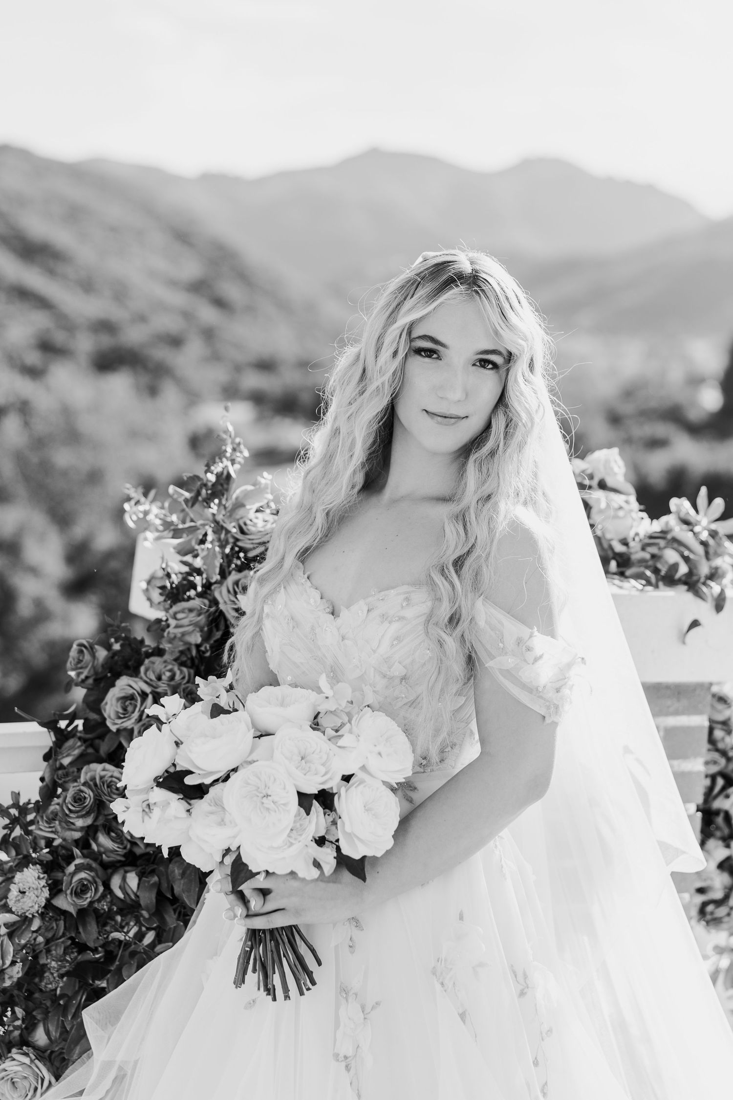 A bride in a white lace gown poses outdoors with a lush floral bouquet against a mountain backdrop.