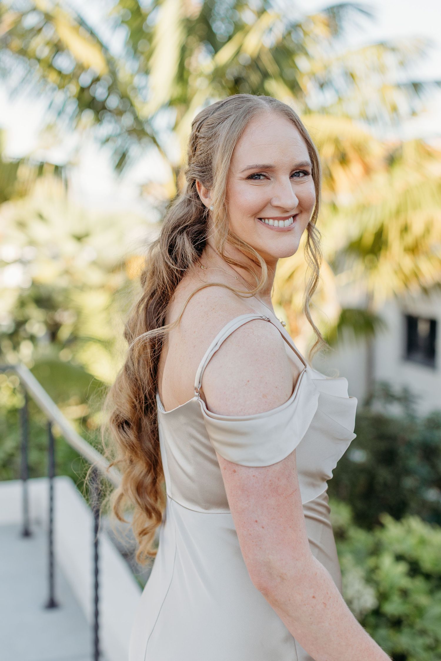 Portrait of person in off-shoulder white dress with long wavy hair against tropical palm background.