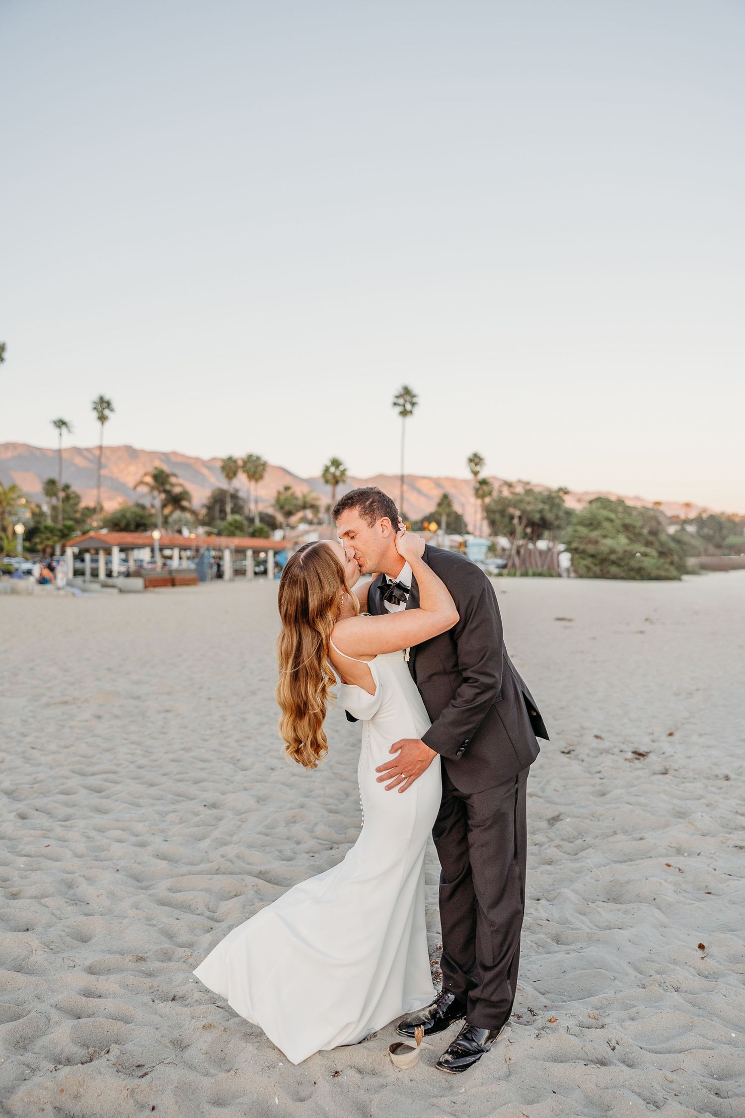 Romantic beach wedding photo of a couple embracing on the sand with palm trees in the background.