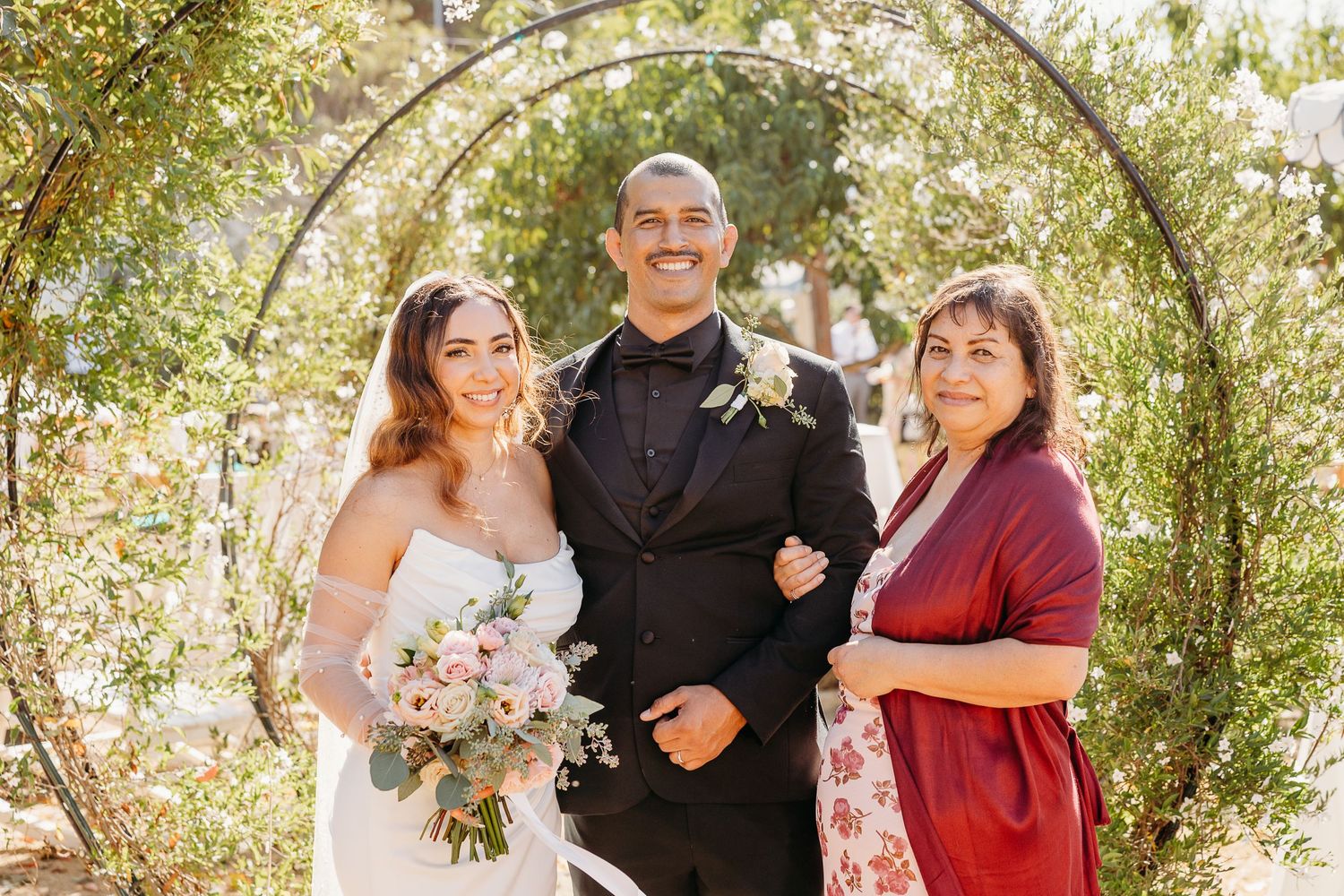Wedding ceremony under a greenery-covered arch with guests seated outdoors.
