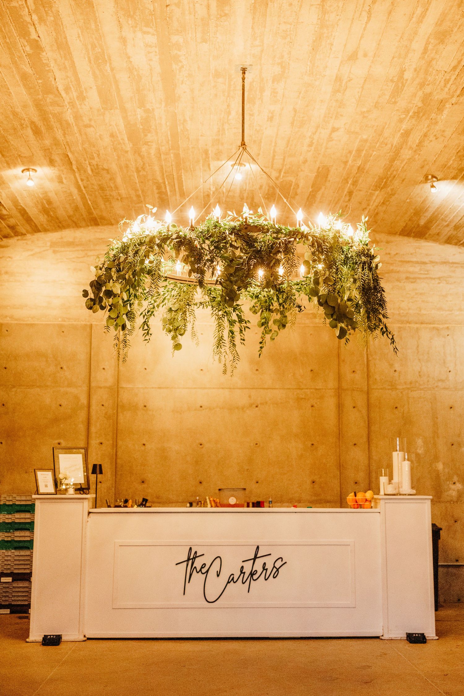 Modern white wedding bar setup with greenery chandeliers and warm lighting in a vaulted ceiling venue.