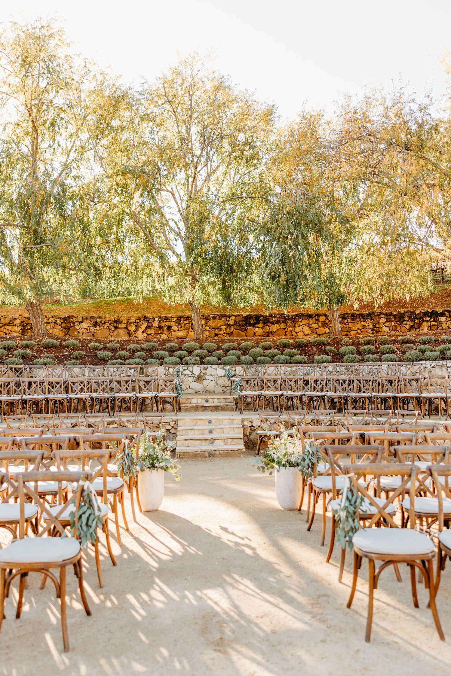 Wooden cross-back chairs arranged in rows for an outdoor wedding ceremony against a stone wall.
