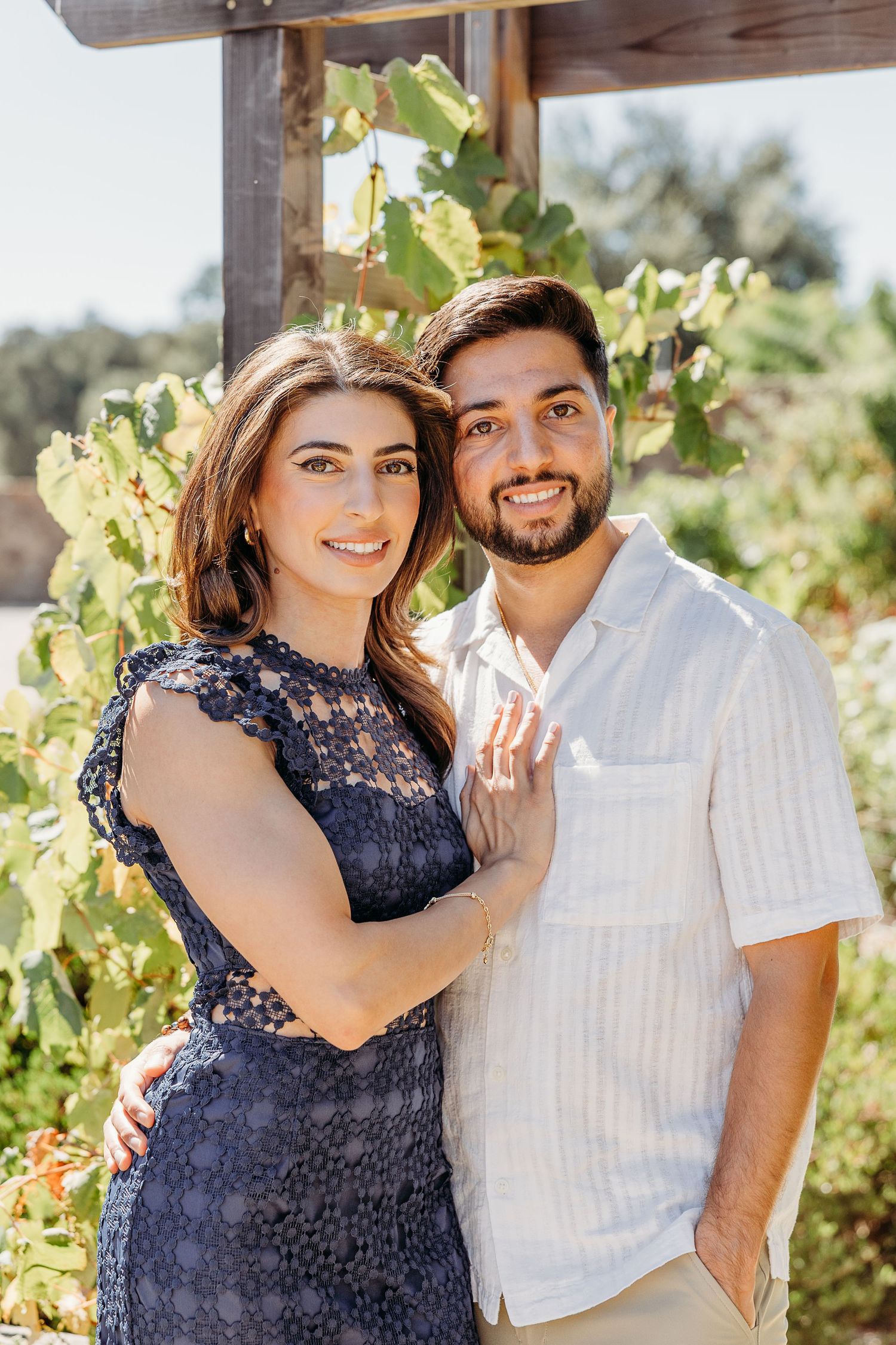 Couple poses together in vineyard wearing navy dress and white shirt.