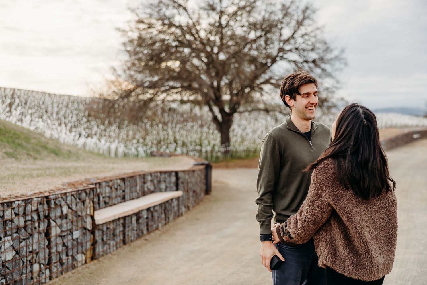 A couple shares an intimate moment together outdoors on a cloudy day with a tree in the background.