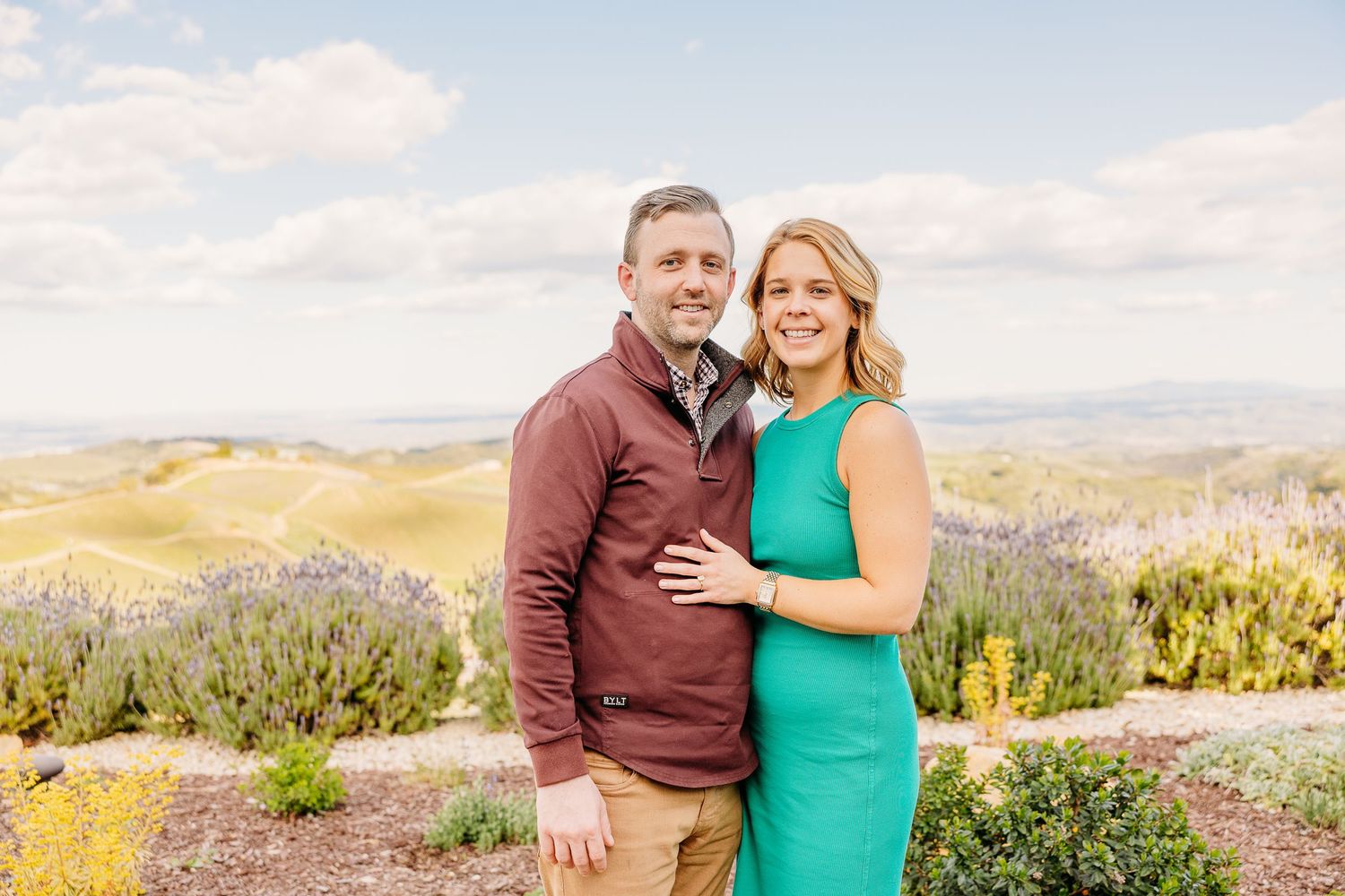A couple in burgundy and green attire embraces while posing for outdoor mountain landscape photos at sunset.