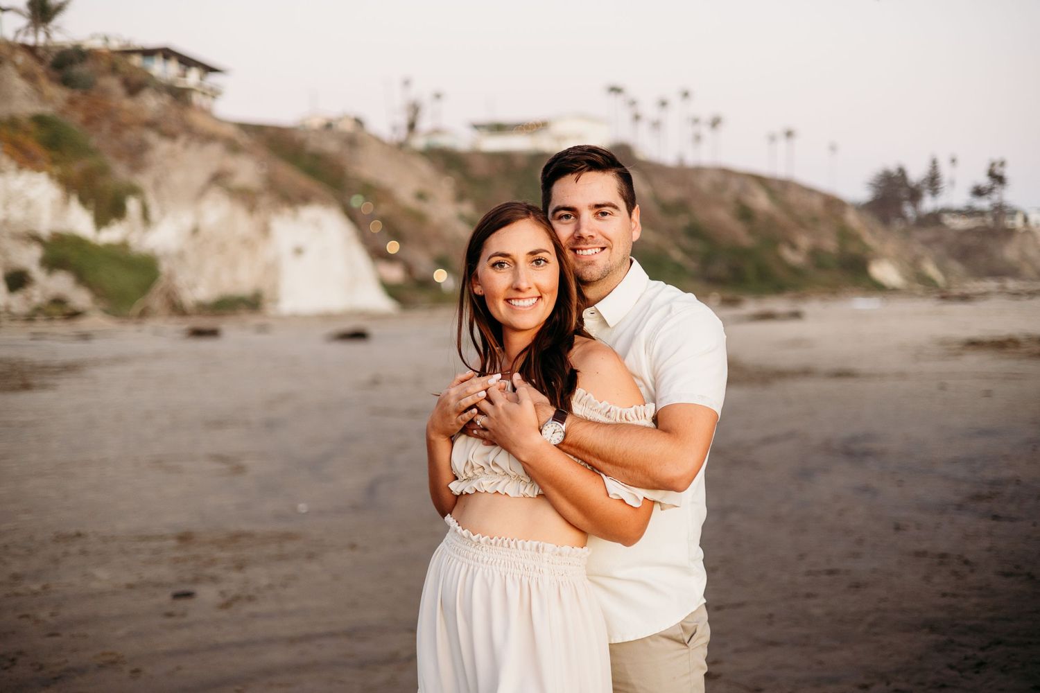 A couple embraces on a beach during sunset with coastal cliffs visible in the background.