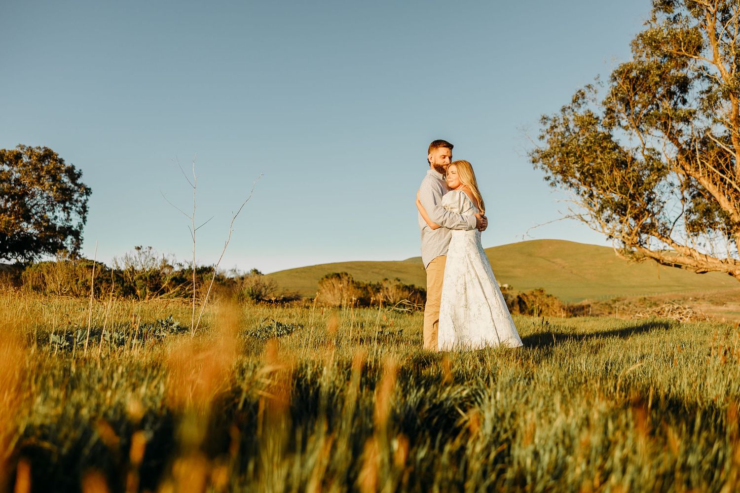 A couple embraces in a golden rural landscape with tall grass and golden hour sunlight during a wedding photoshoot.