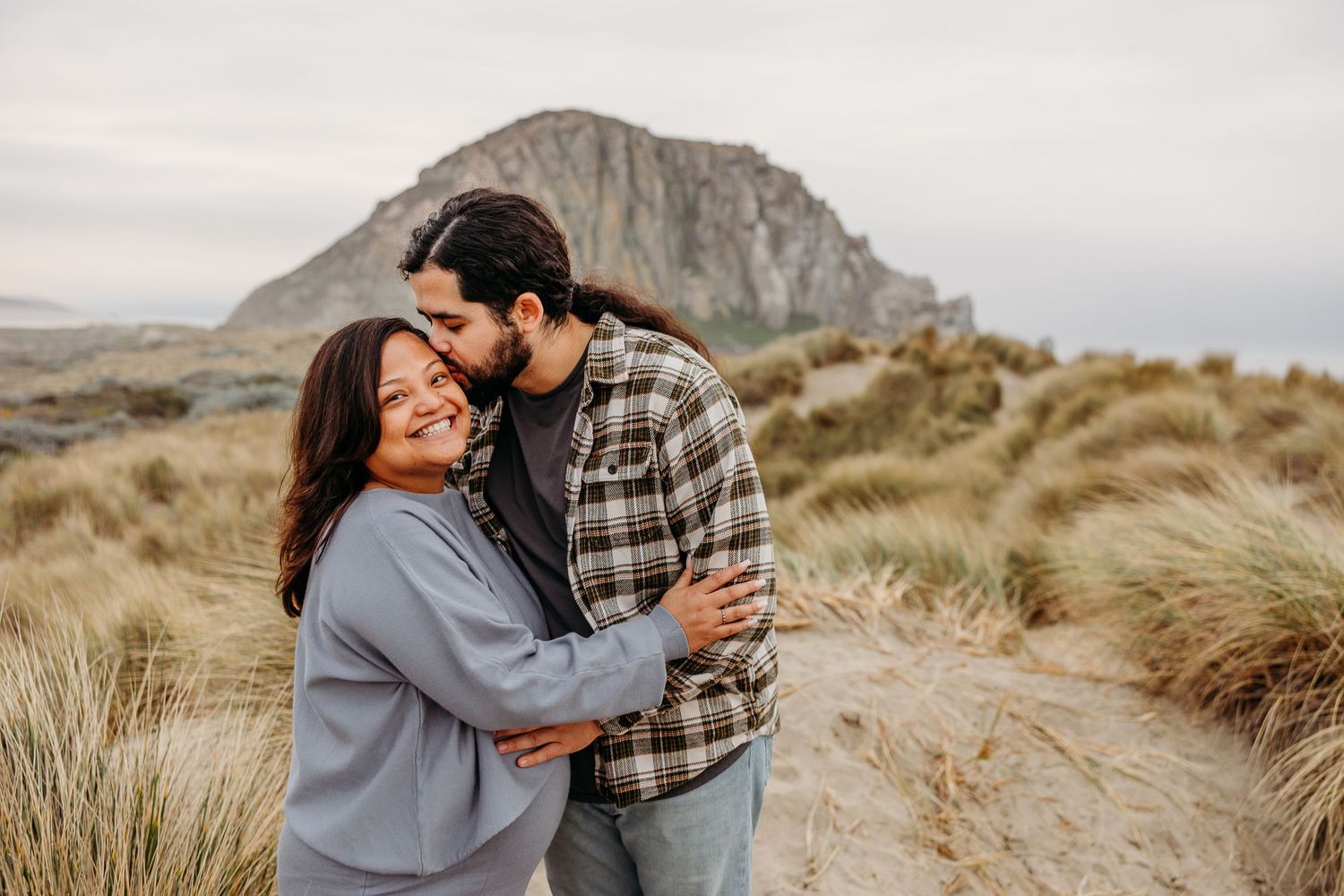 Romantic couple embracing on a grassy coastal path with a dramatic rocky mountain backdrop.