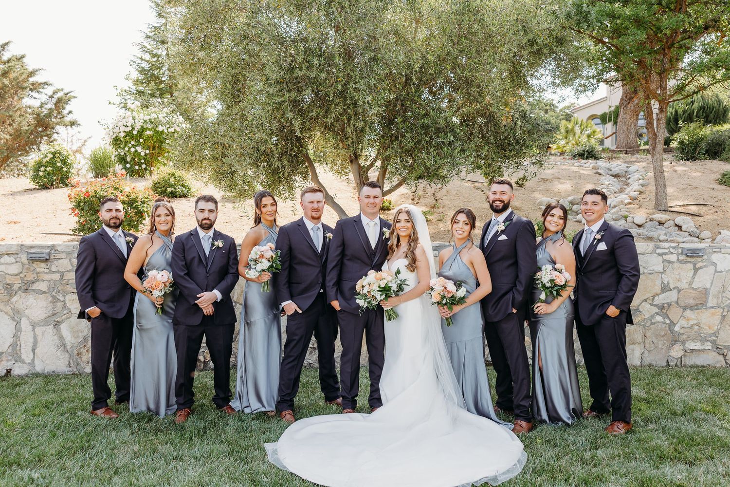 Wedding party poses outdoors against stone wall with bridesmaids in gray dresses and groomsmen in dark suits.