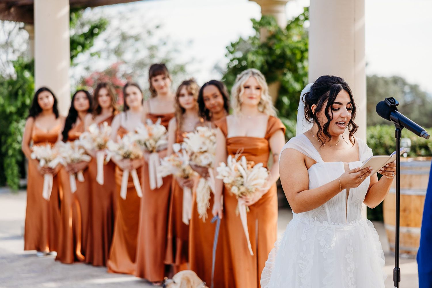 Bridesmaids in burnt orange dresses watch as bride delivers speech at outdoor wedding venue.