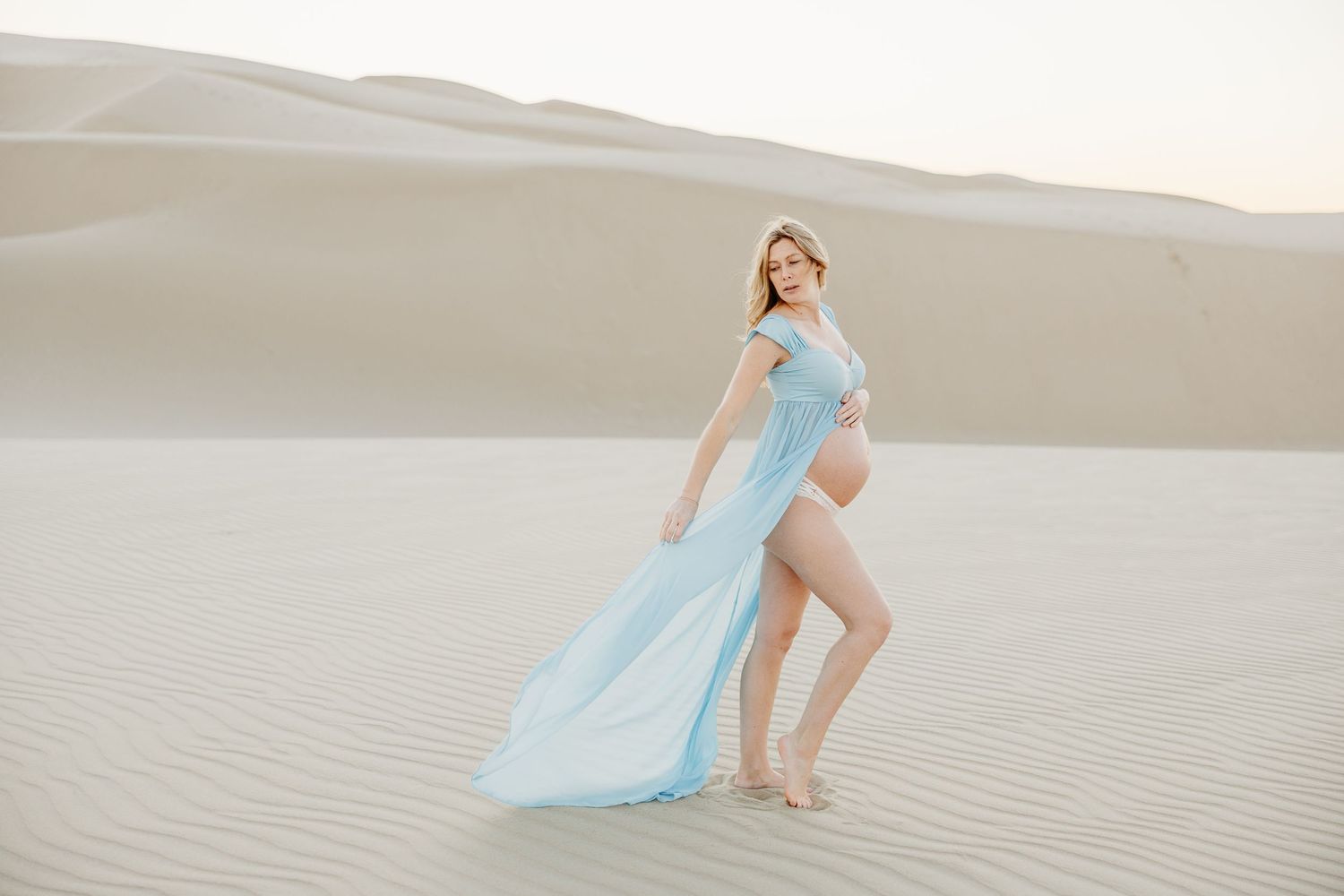 Dreamy maternity portrait with flowing light blue dress in desert landscape.