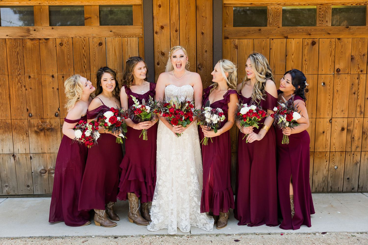 Wedding party in burgundy dresses poses against rustic wooden doors with red bouquets.
