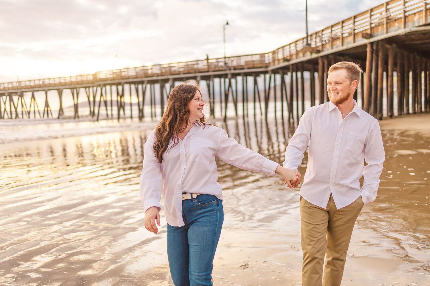 Two people hold hands while walking barefoot on wet sand near a beach pier at sunset.