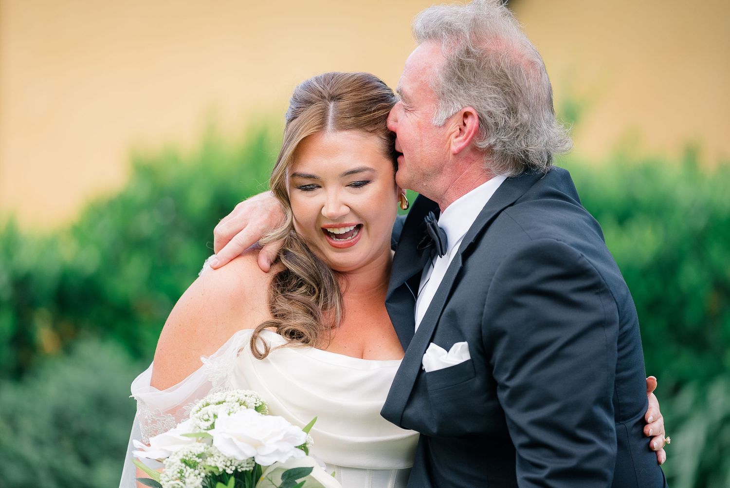 Bride laughing joyfully as older man in tuxedo kisses her cheek.