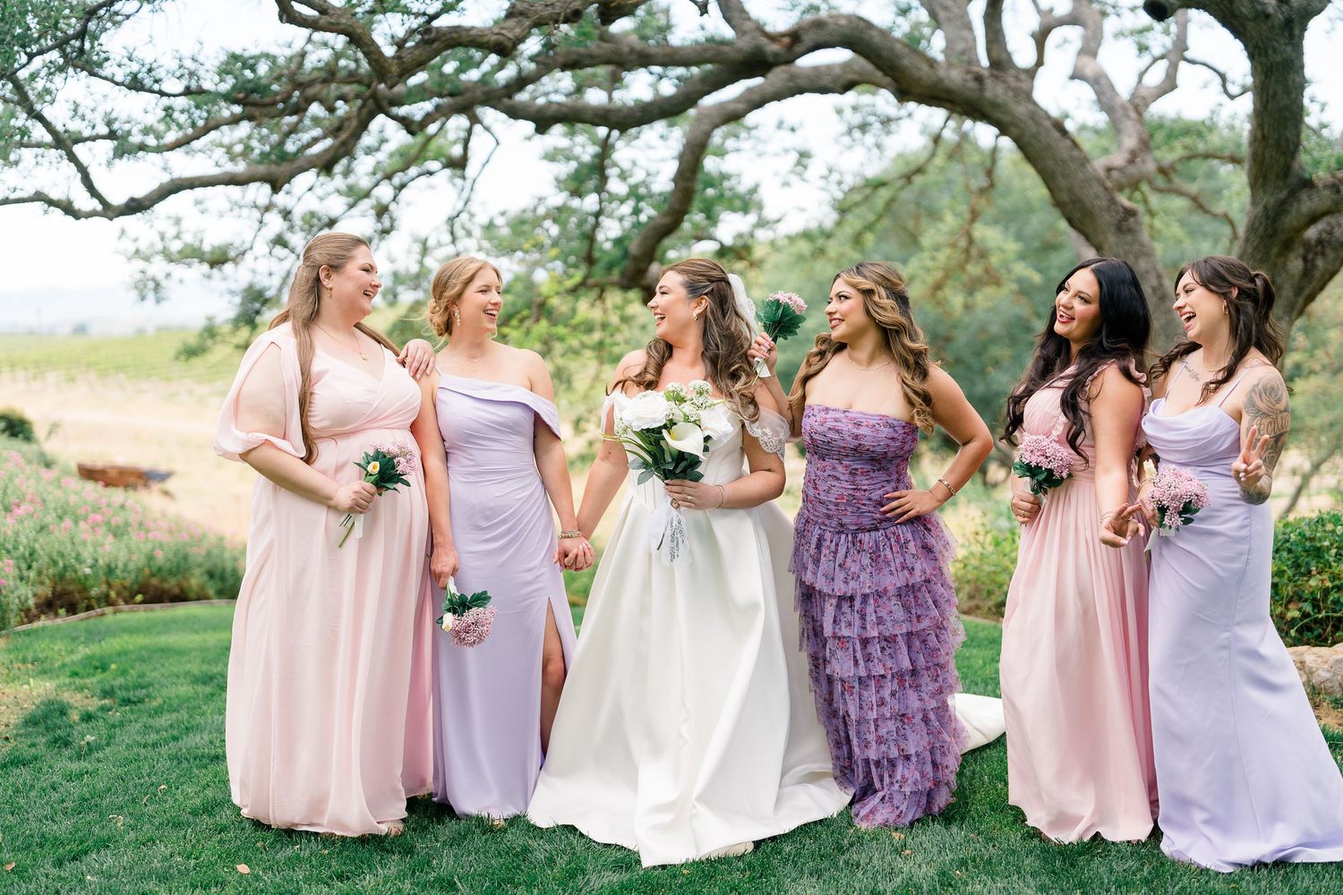 Bride with bridesmaids in pink and lavender gowns laughing outdoors.