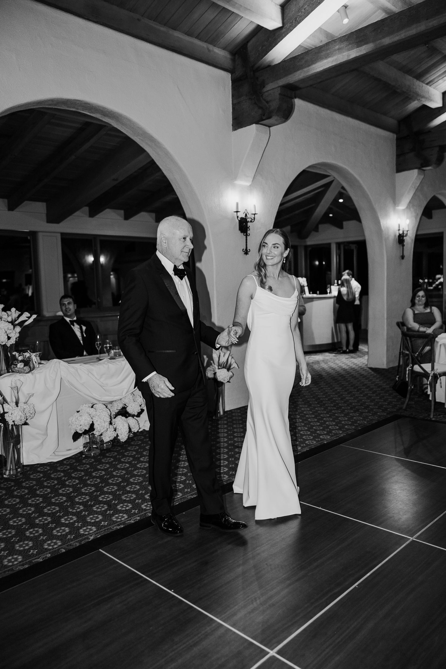 Black and white photo of a couple dancing in an elegant reception venue with arched ceilings.