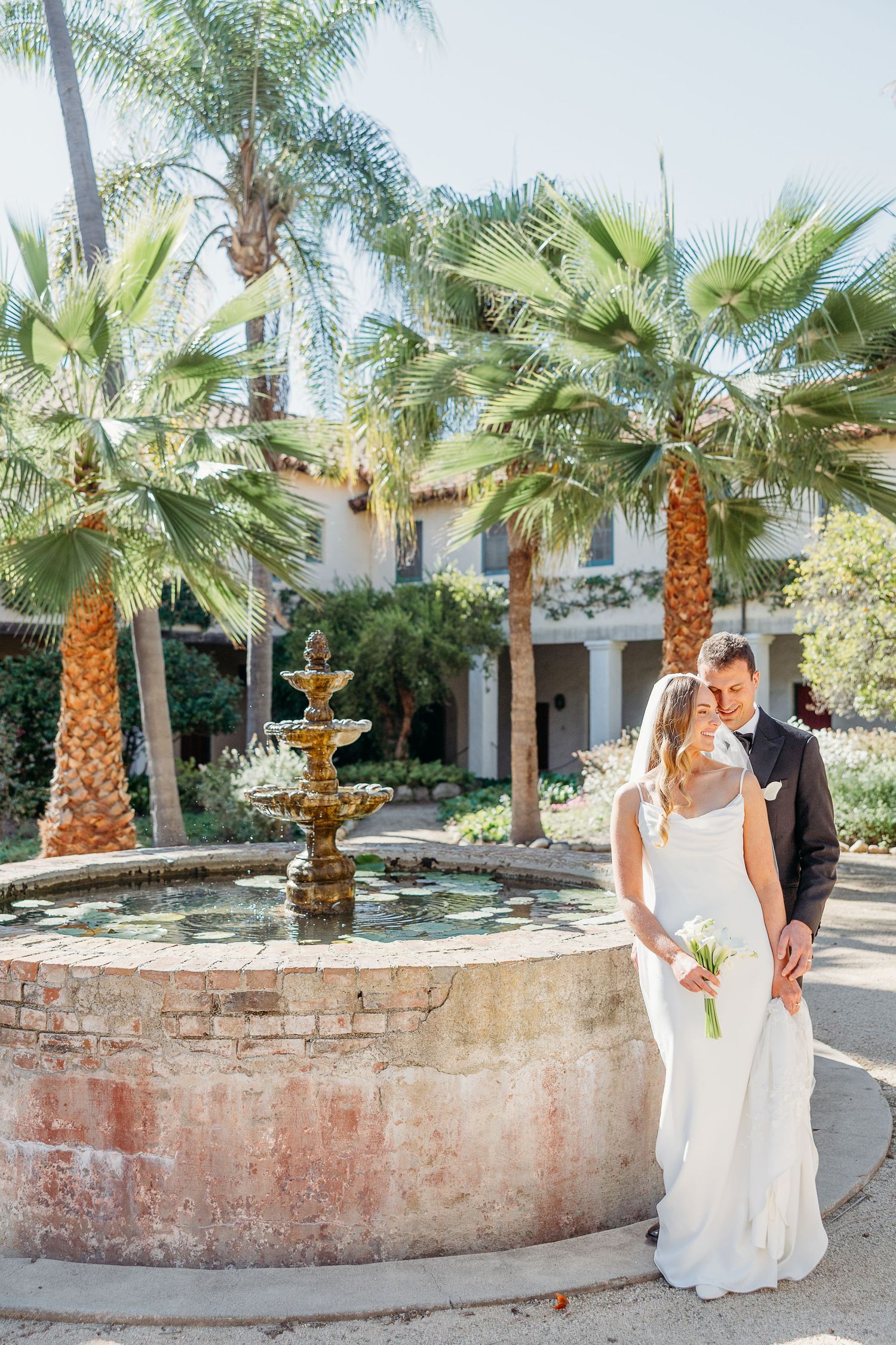 A couple shares an intimate moment in a tropical garden setting with palm trees and stone walls.