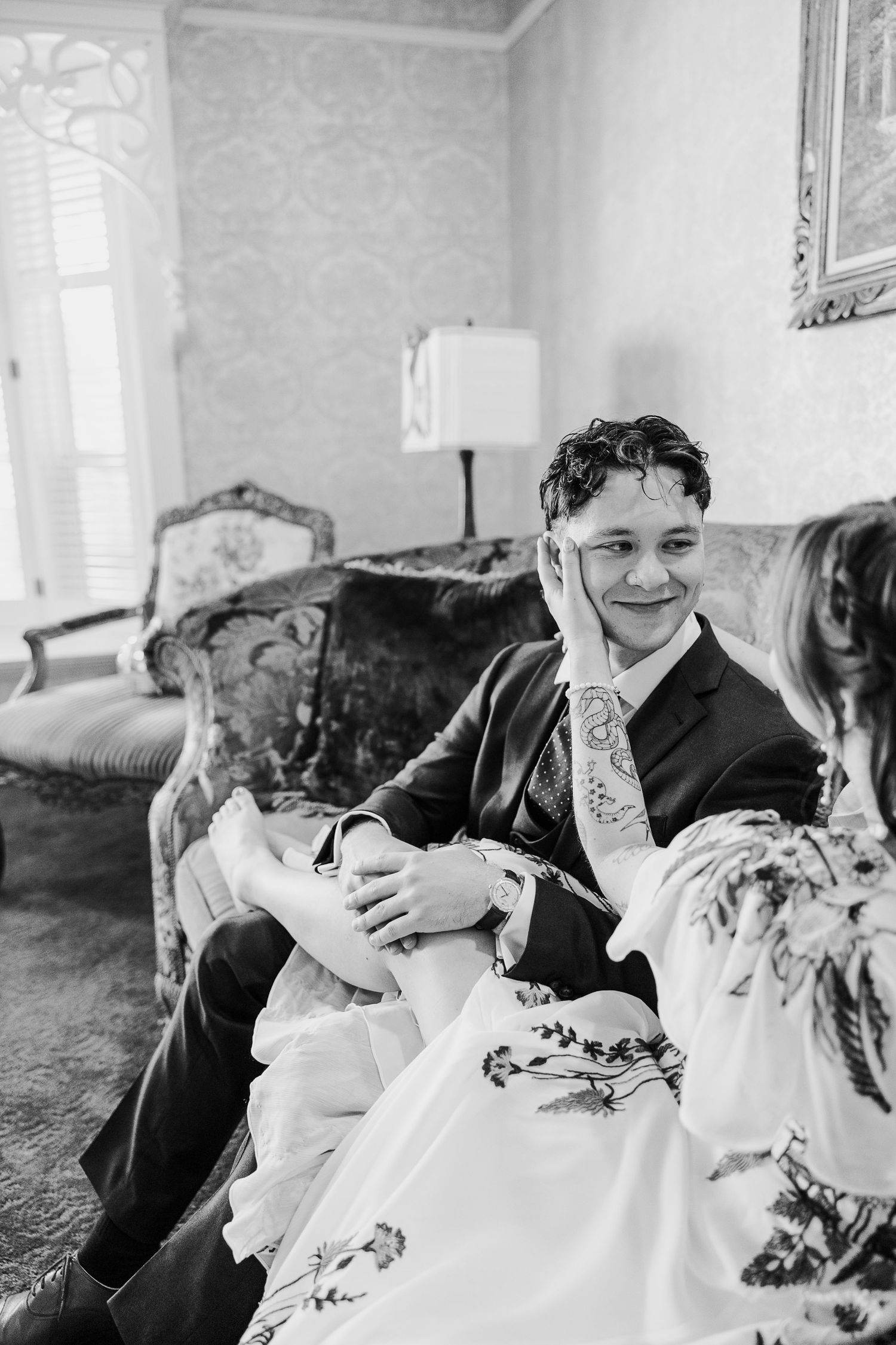 A candid black and white photo of a couple relaxing on an elegant vintage couch with a table lamp in the background.