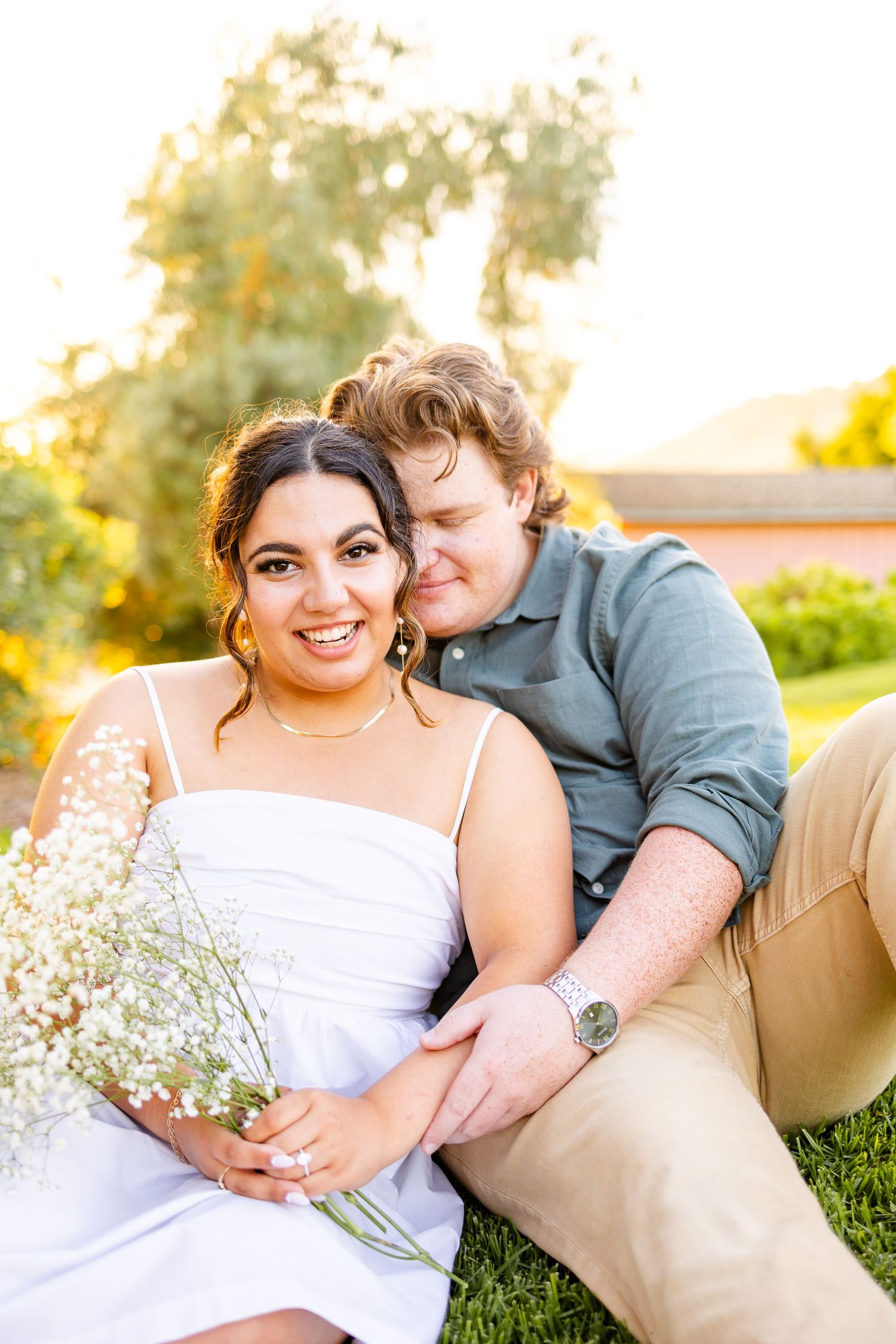 Couple sharing tender moment on blanket with delicate white flowers in sunlit field.