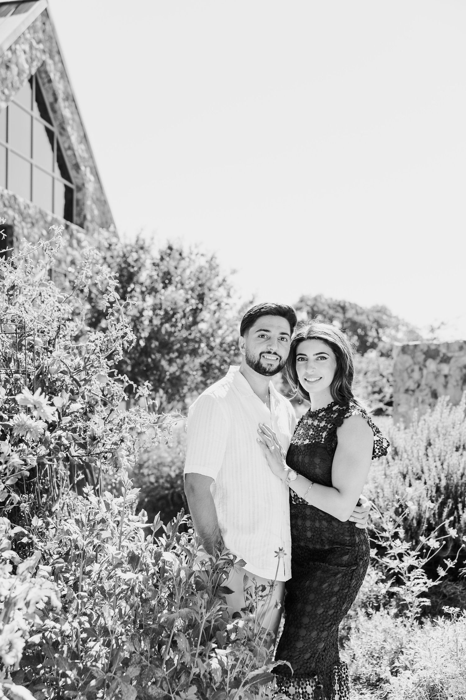 A couple laughs together in a sunny garden filled with blooming flowers.