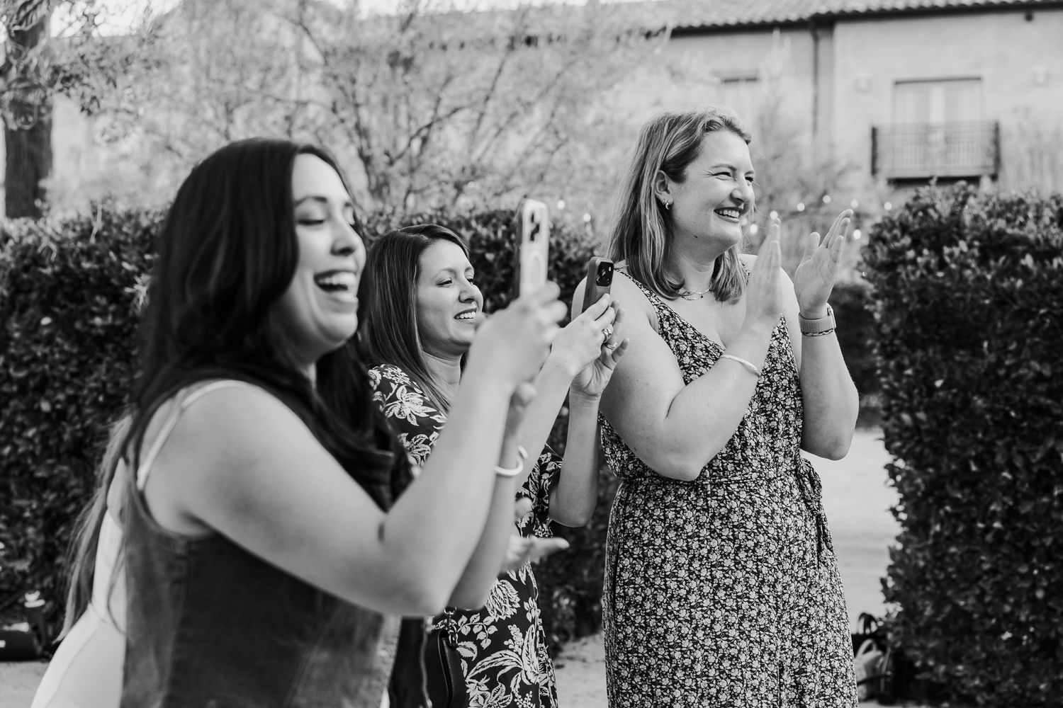 A candid black and white photo shows three people laughing joyfully together in a garden setting.