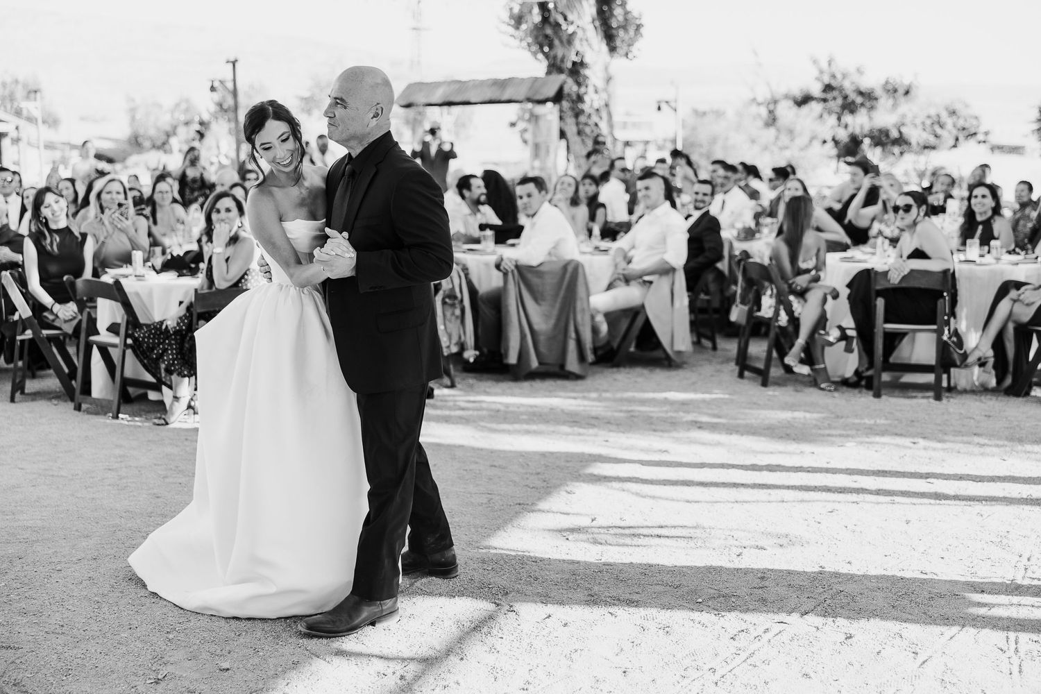 Wedding celebration scene with guests seated at outdoor reception tables under string lights.
