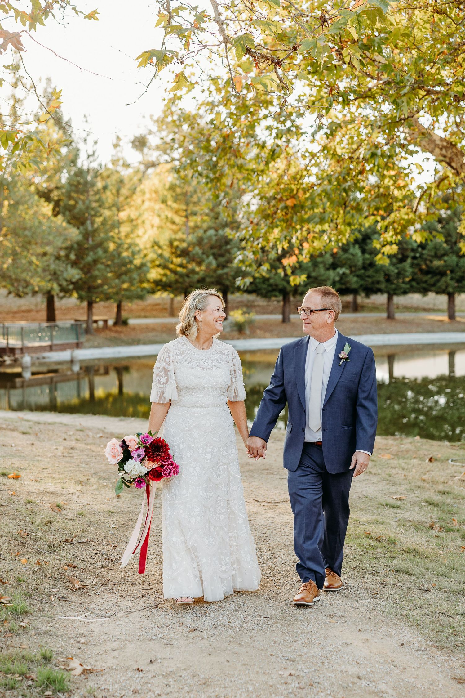 A couple walks together in a sunlit autumn orchard with golden leaves and warm lighting.