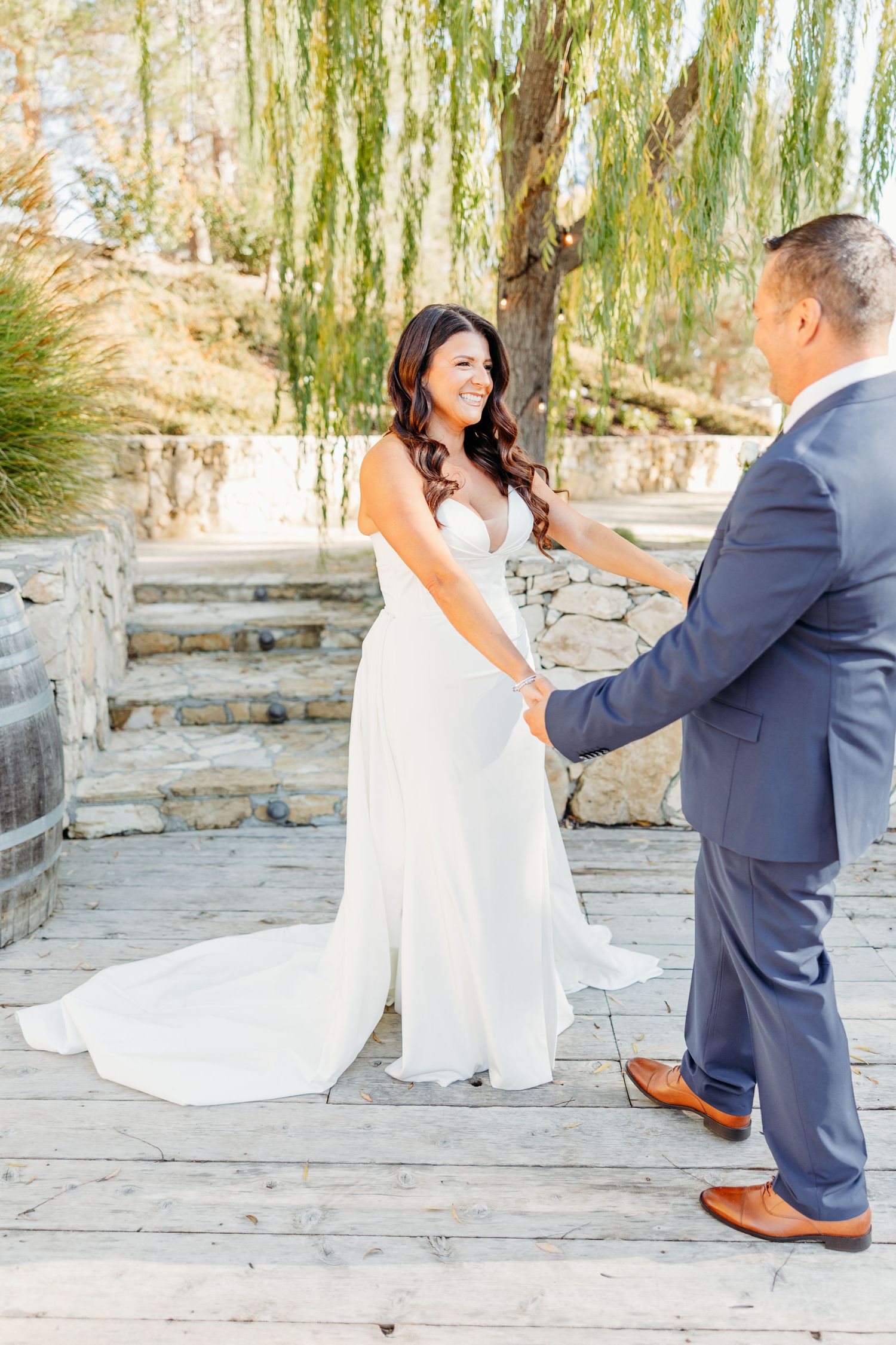 A playful wedding couple dances together on a stone patio with willow trees in the background.