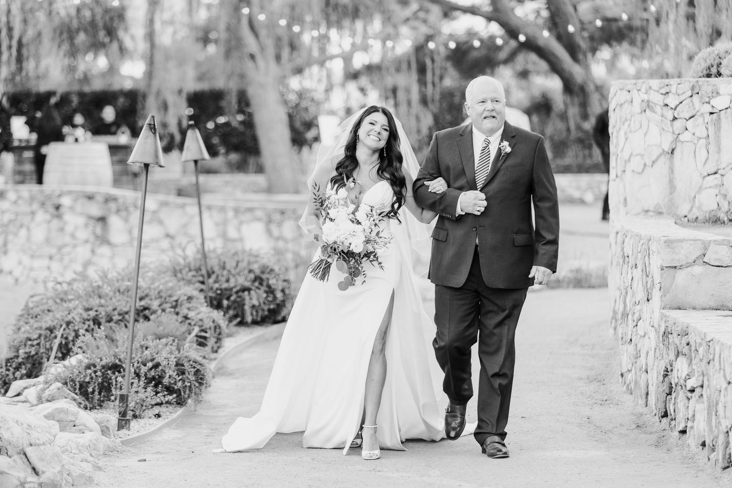 Wedding couple walks hand in hand along stone pathway surrounded by natural garden scenery.