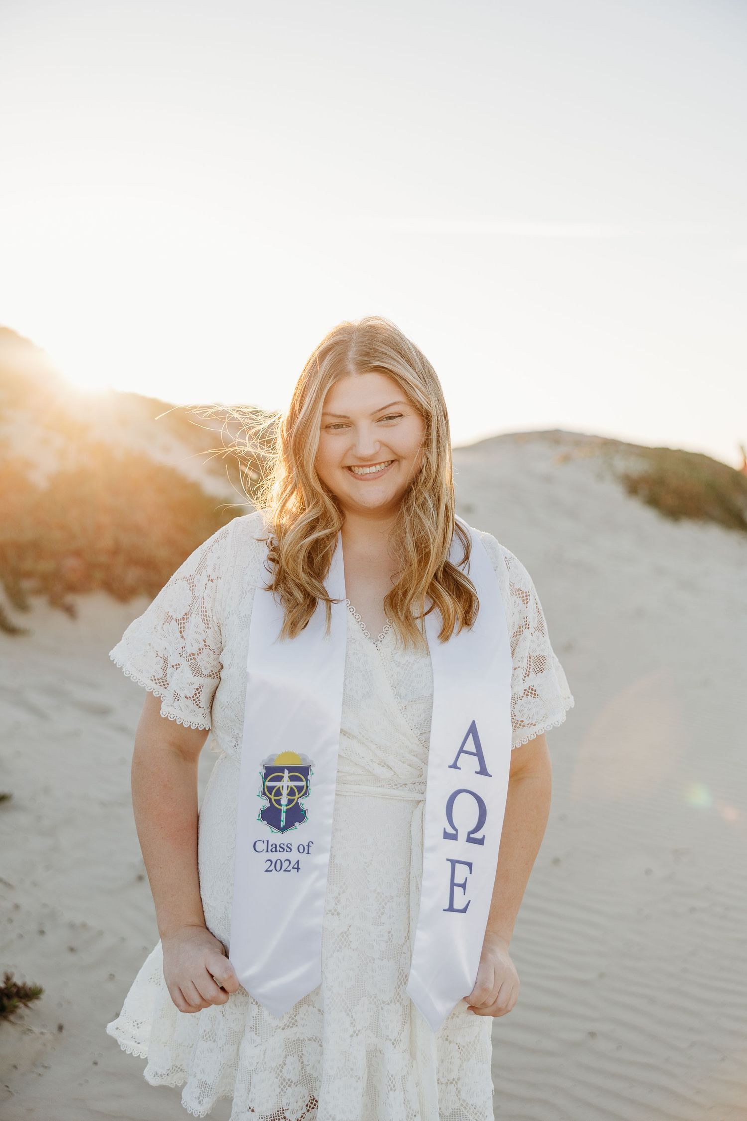 Graduate wearing white dress and sorority sash poses in front of sandy dunes at sunset.