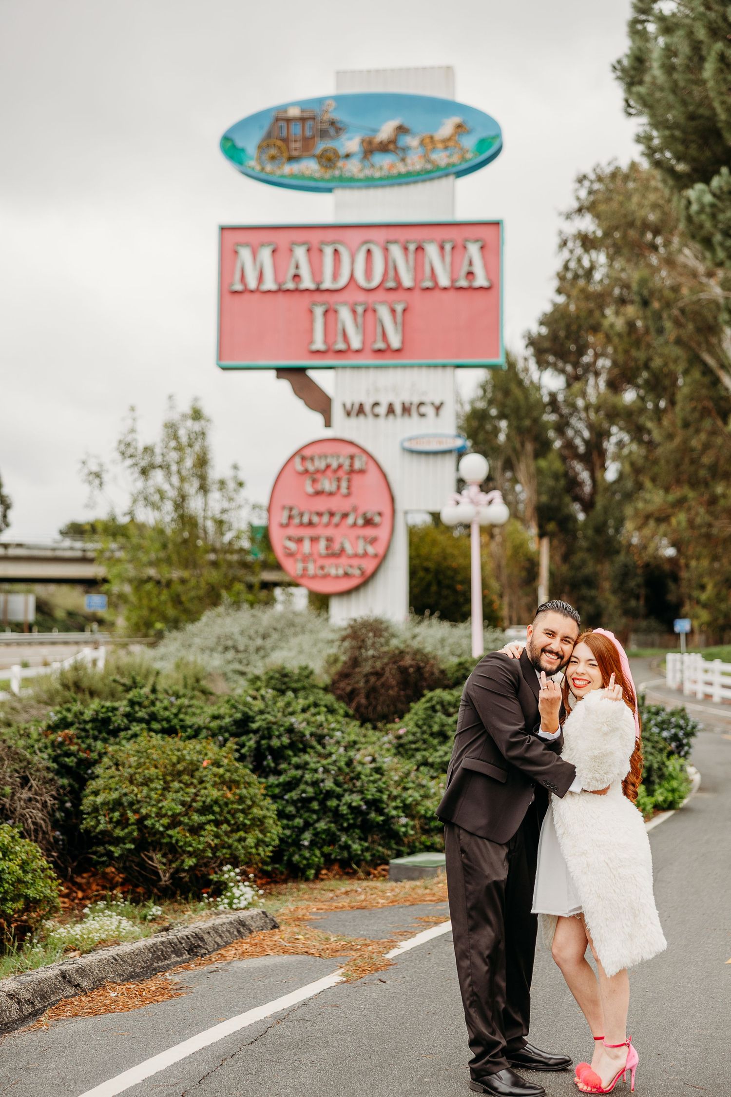A couple embraces in front of the retro pink Madonna Inn sign with vintage styling and pink accents.